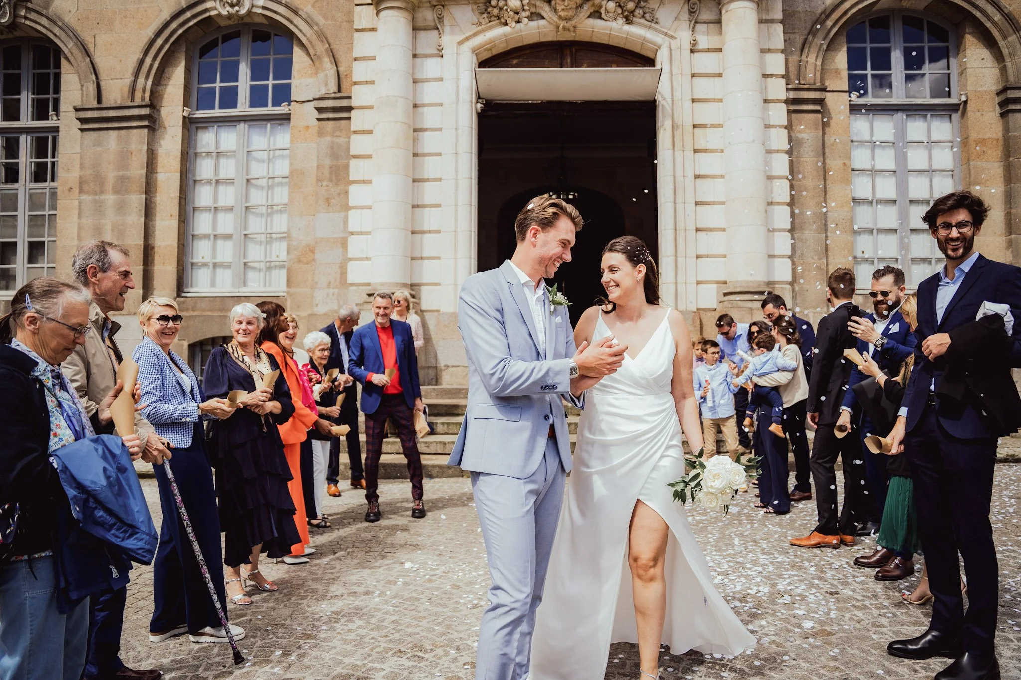 Un couple de mariés, un homme en costume gris clair et une femme en robe blanche, marche main dans la main devant une foule d'invités lors d'un mariage en plein air devant un bâtiment en pierre ancienne, avec des invités qui sourient et lancent des c