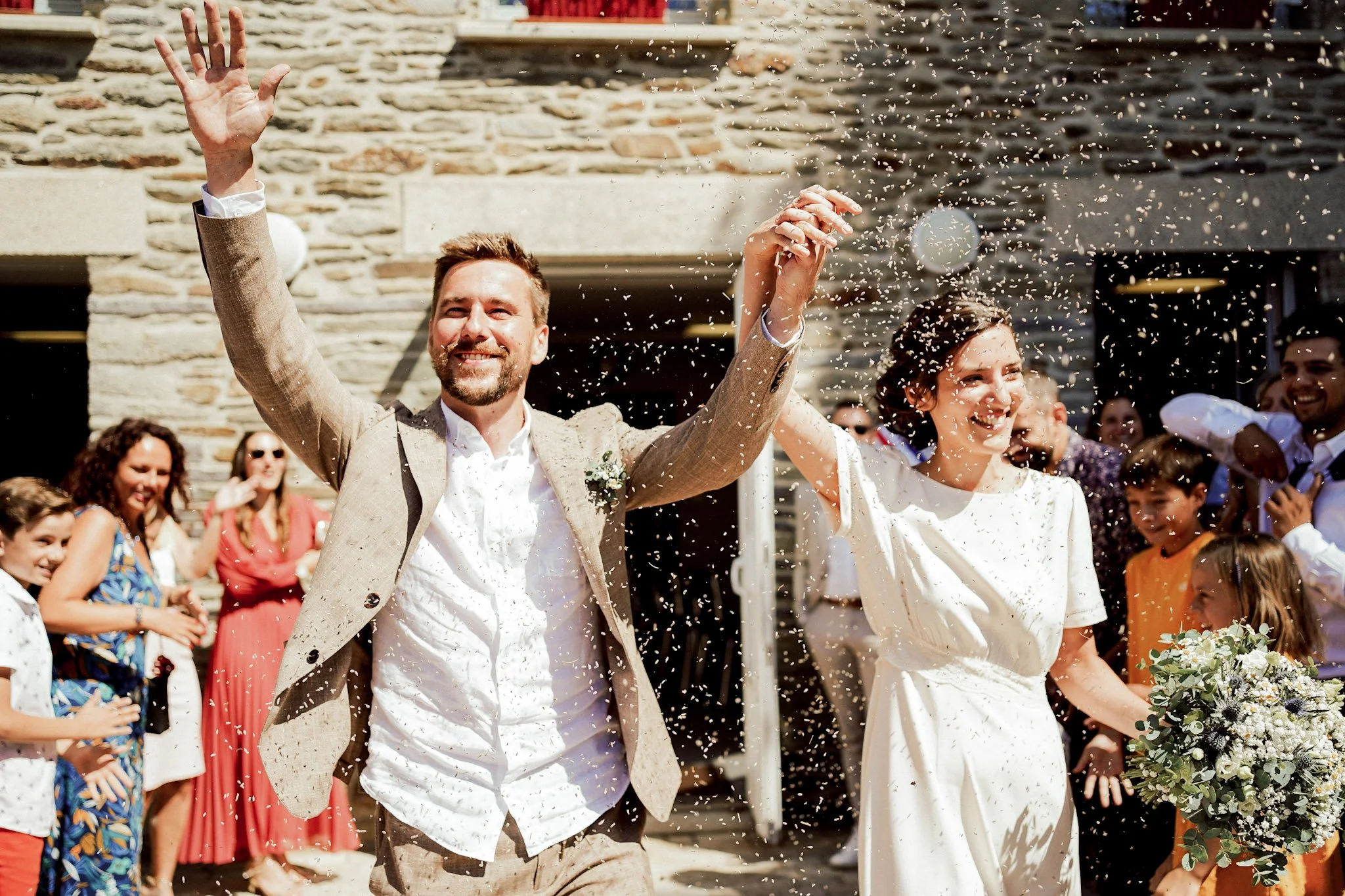 Un couple de mariés heureux, main dans la main, lors d'une célébration en plein air, entouré d'invités ensoleillés, avec un fond en pierre, célébrant leur mariage avec de la confettis