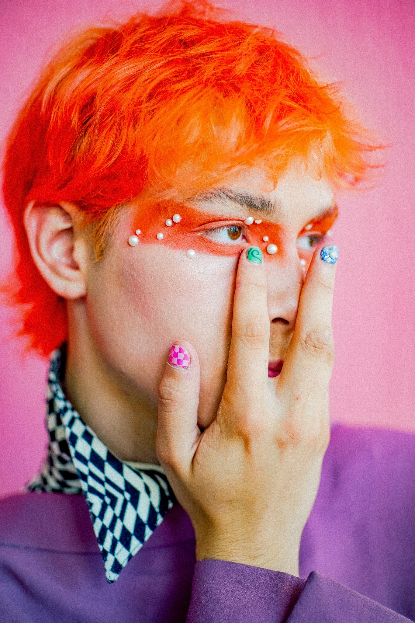 Un portrait d'une personne avec des cheveux orange, maquillage audacieux avec des détails en perles autour des yeux et ongles colorés, portant une chemise à carreaux sous un vêtement violet, devant un fond rose.