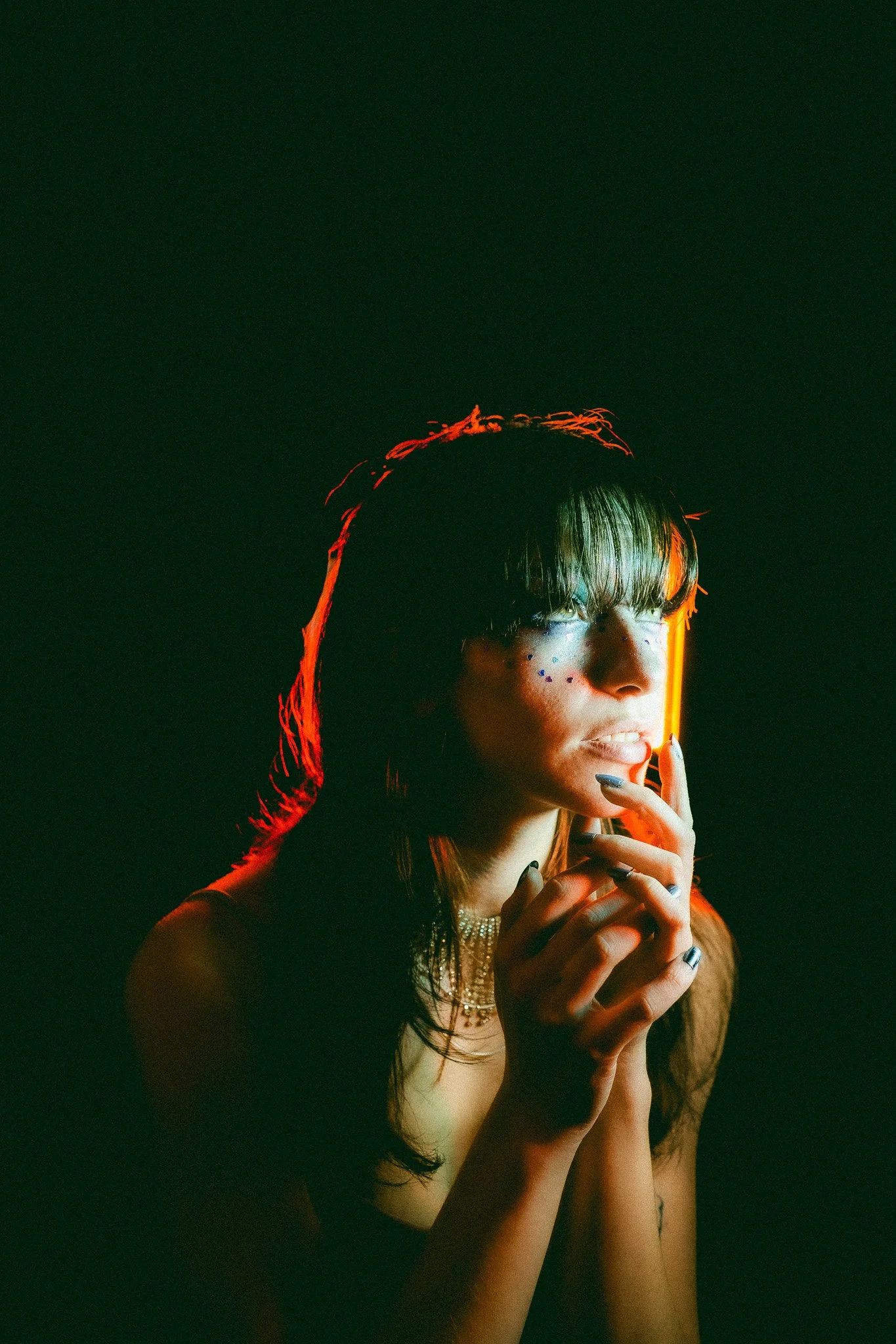 Portrait d'une femme avec maquillage artistique, portant un collier de perles, tenant une cigarette, éclairée par une lumière rouge-orange sur un fond sombre.
