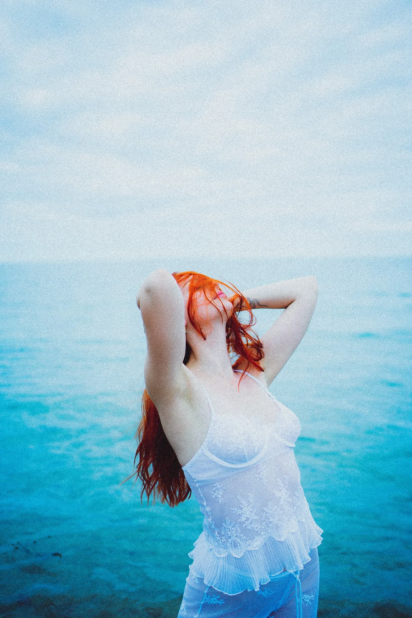 Femme aux cheveux rouges posant devant la mer, portant une robe en dentelle blanche.