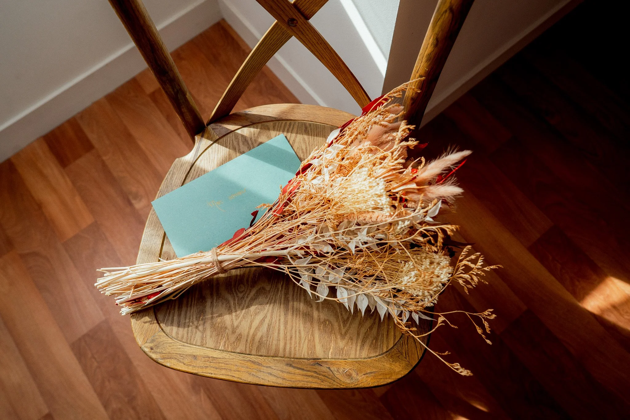 Un bouquet de fleurs séchées posé sur une chaise en bois, avec une carte bleue. La chaise est en bois clair et est située sur un sol en parquet.