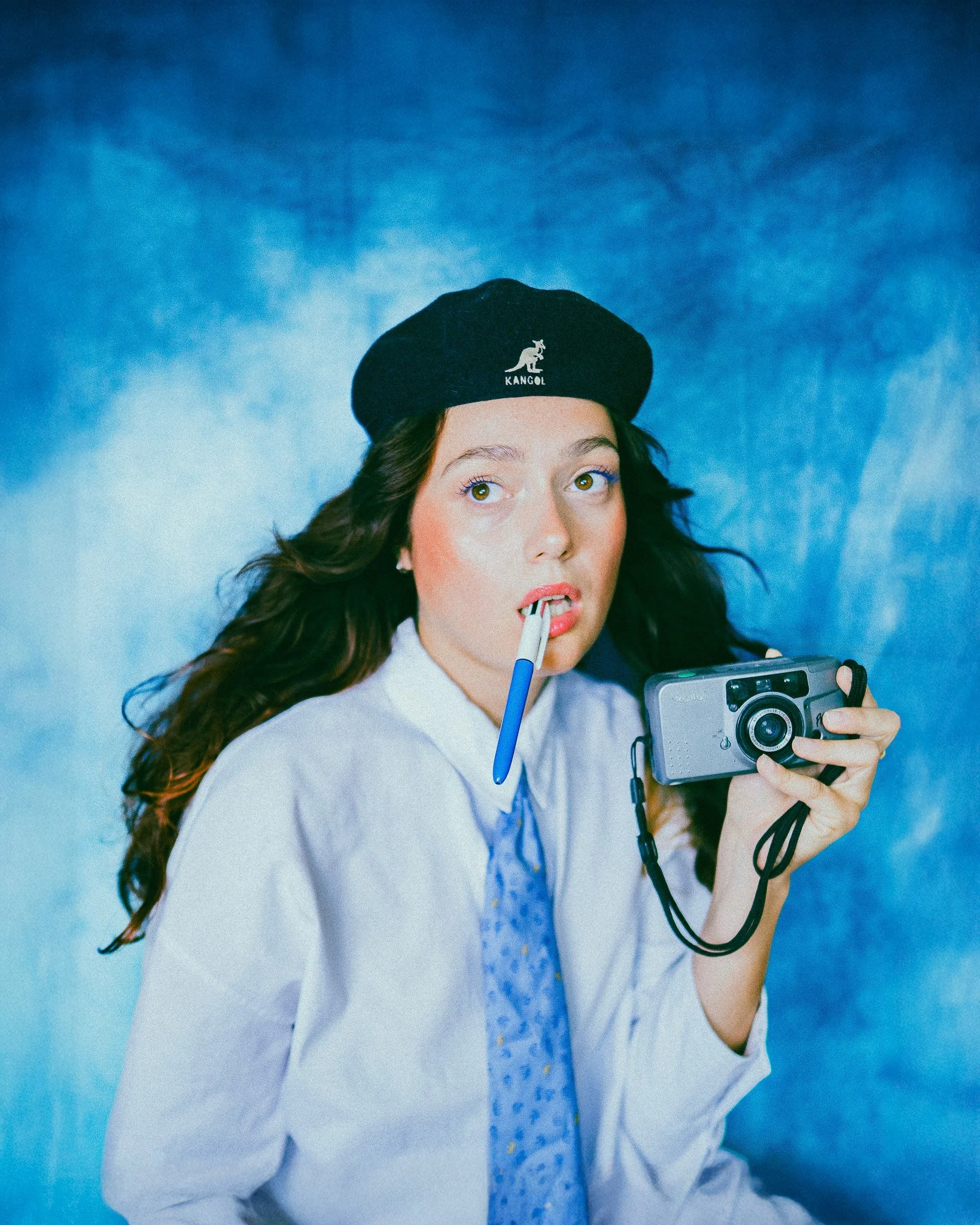 Femme avec un béret noir, tenue de bureau, tenant un appareil photo, regardant vers l'objectif, avec un stylo dans la bouche, fond bleu.