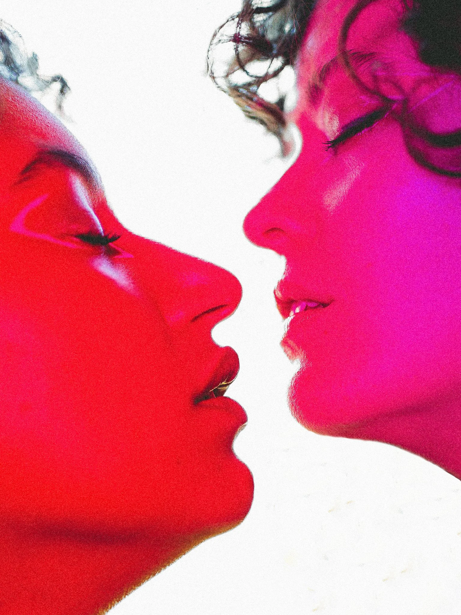 Deux femmes se font face, yeux fermés, avec des lèvres presque touchantes, l'une en maquillage rouge vif et l'autre en maquillage rose, création artistique avec jeux de lumière.