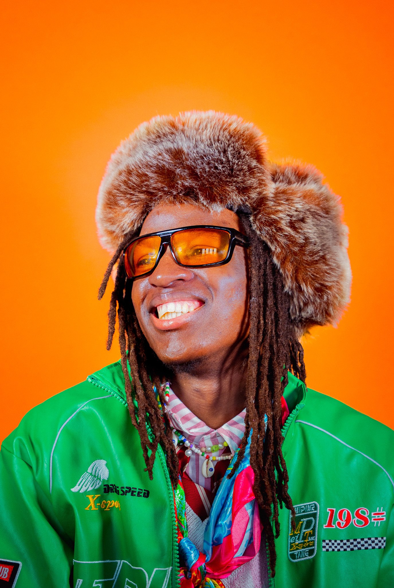 Un homme souriant portant un chapeau en fourrure, des lunettes orange et une veste verte contre un fond orange.