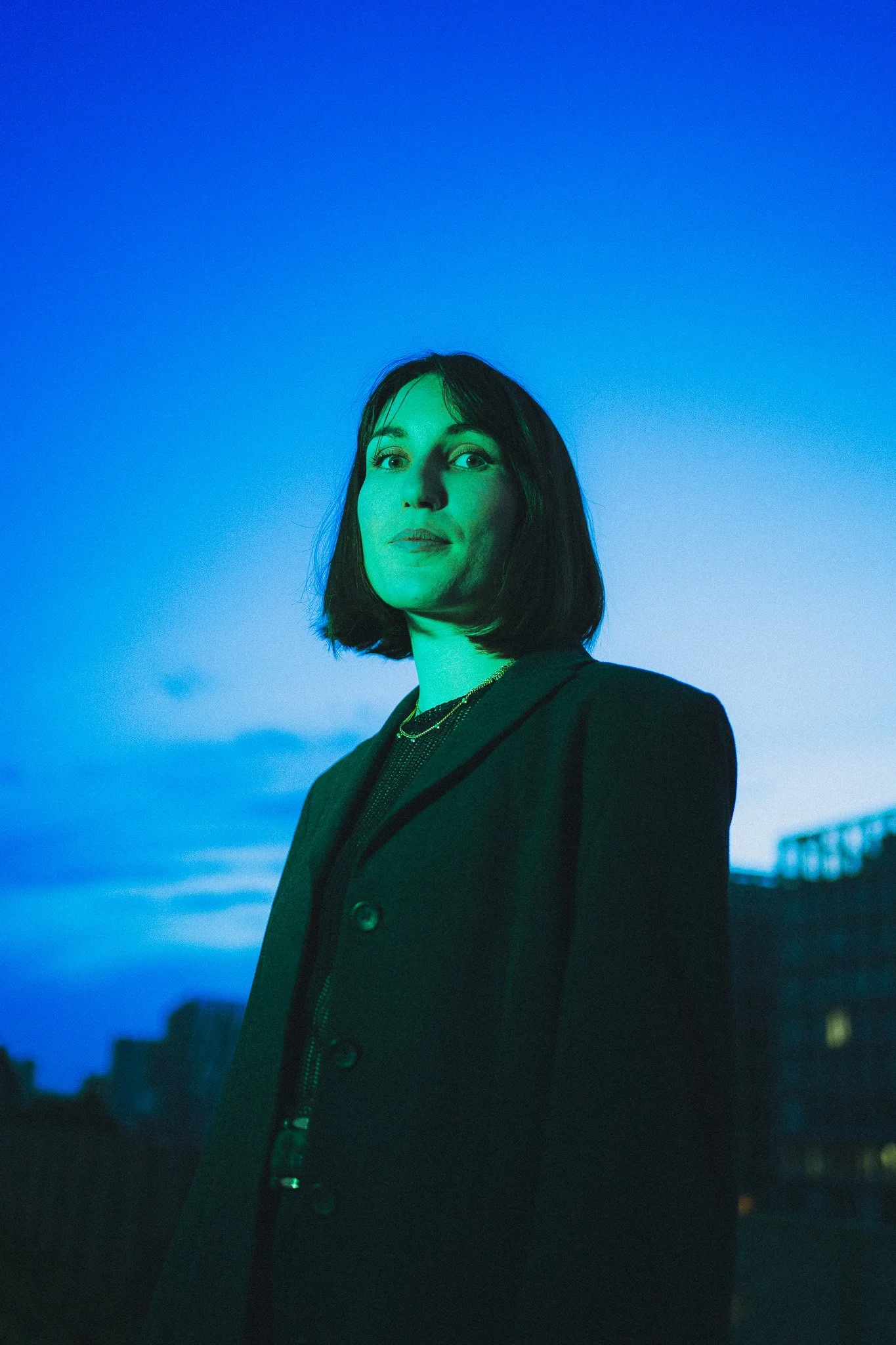 Une femme avec une coupe au carré, habillée en blazer noir, se tient à l'extérieur au crépuscule avec un ciel bleu en arrière-plan.