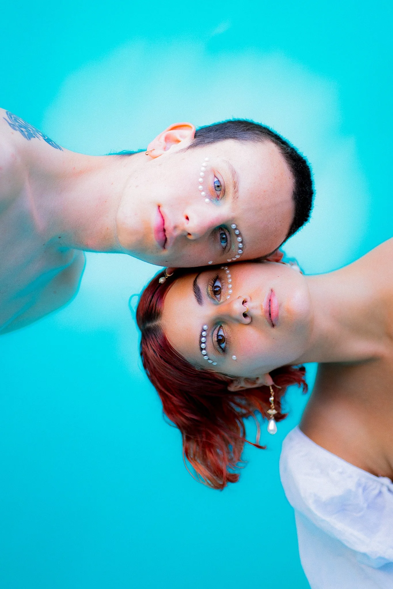 Deux jeunes personnes, un homme et une femme, posent sous l'eau avec une lumière bleue, portant des bijoux et du maquillage décoratif sur le visage.