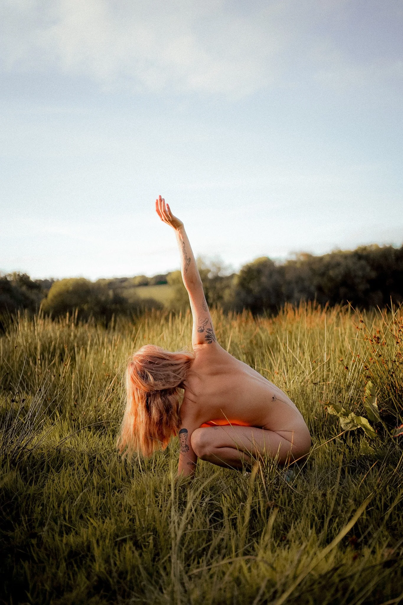 Femme nue faisant une pose de yoga dans un champ d'herbes hautes, avec un ciel clair en arrière-plan.