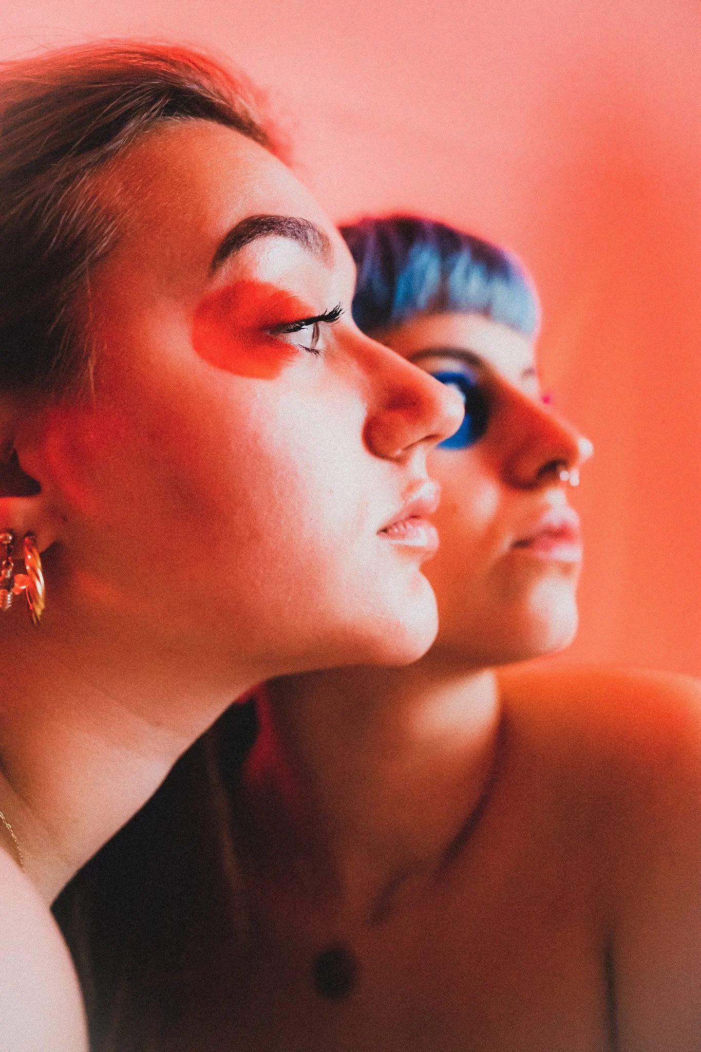 Deux femmes avec des maquillages colorés, regardant vers la droite, avec un arrière-plan rose.