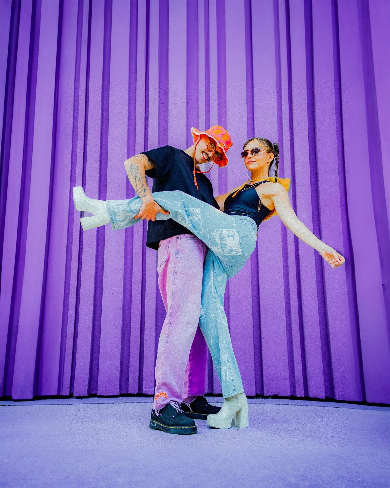 Deux personnes posent devant un mur violet, la personne à gauche porte un chapeau orange et une paire de lunettes, et soutient une femme qui porte des chaussures à talons et un haut noir, la femme a une tresse et porte des lunettes de soleil.