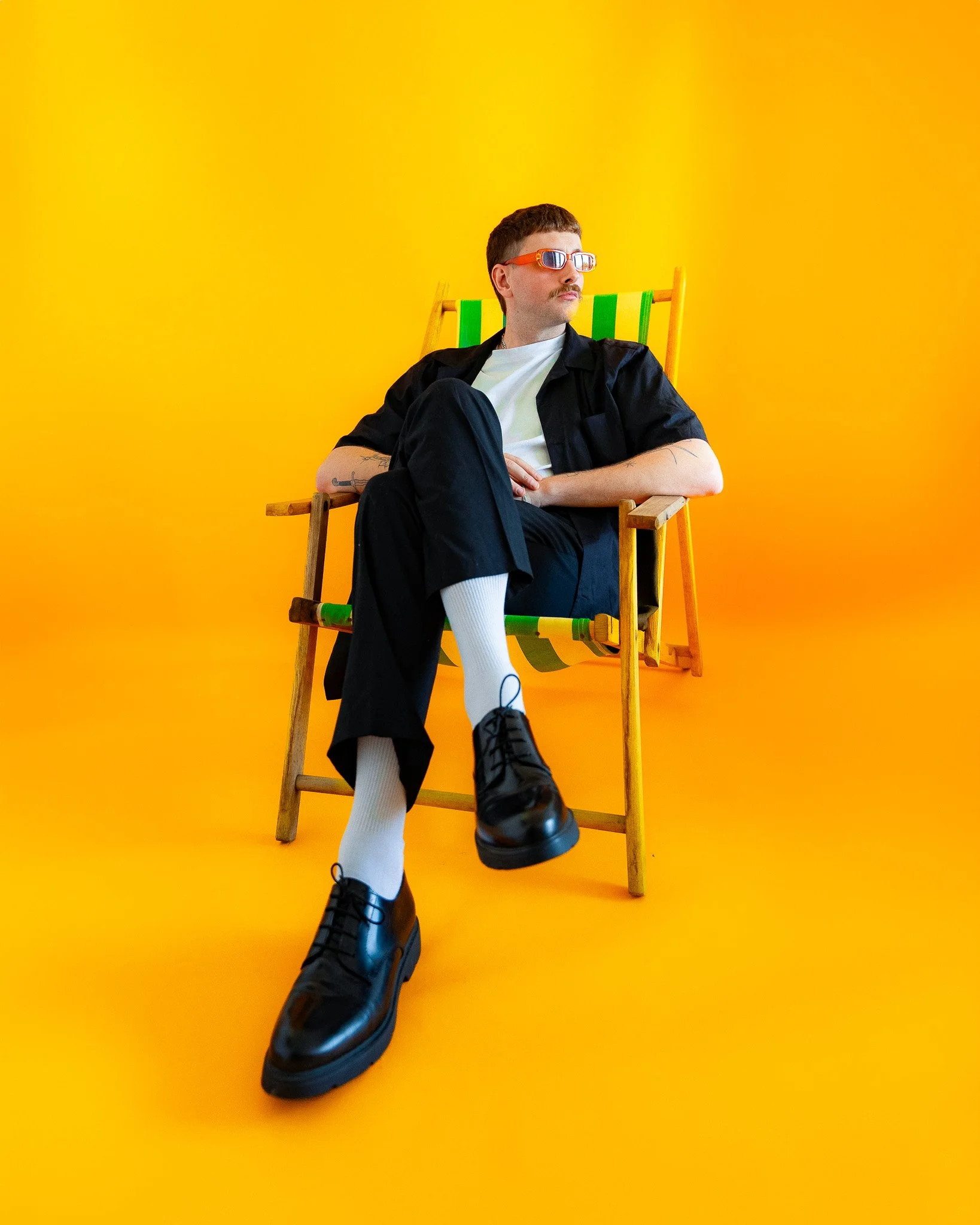 Homme assis dans une chaise en bois sur fond jaune, portant des lunettes de soleil, une chemise noire, un pantalon noir, des chaussettes blanches et des chaussures noires.