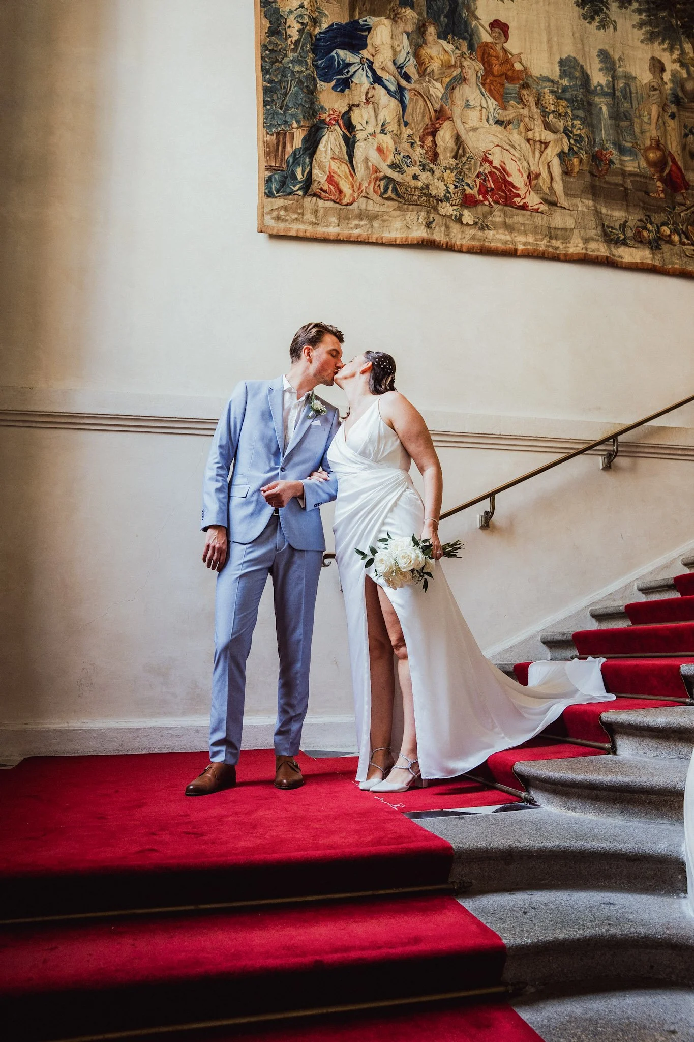 Un couple de mariés s'embrassant en haut d'un escalier recouvert d'un tapis rouge, dans une salle ornée d'une tapisserie ancienne sur le mur derrière eux.