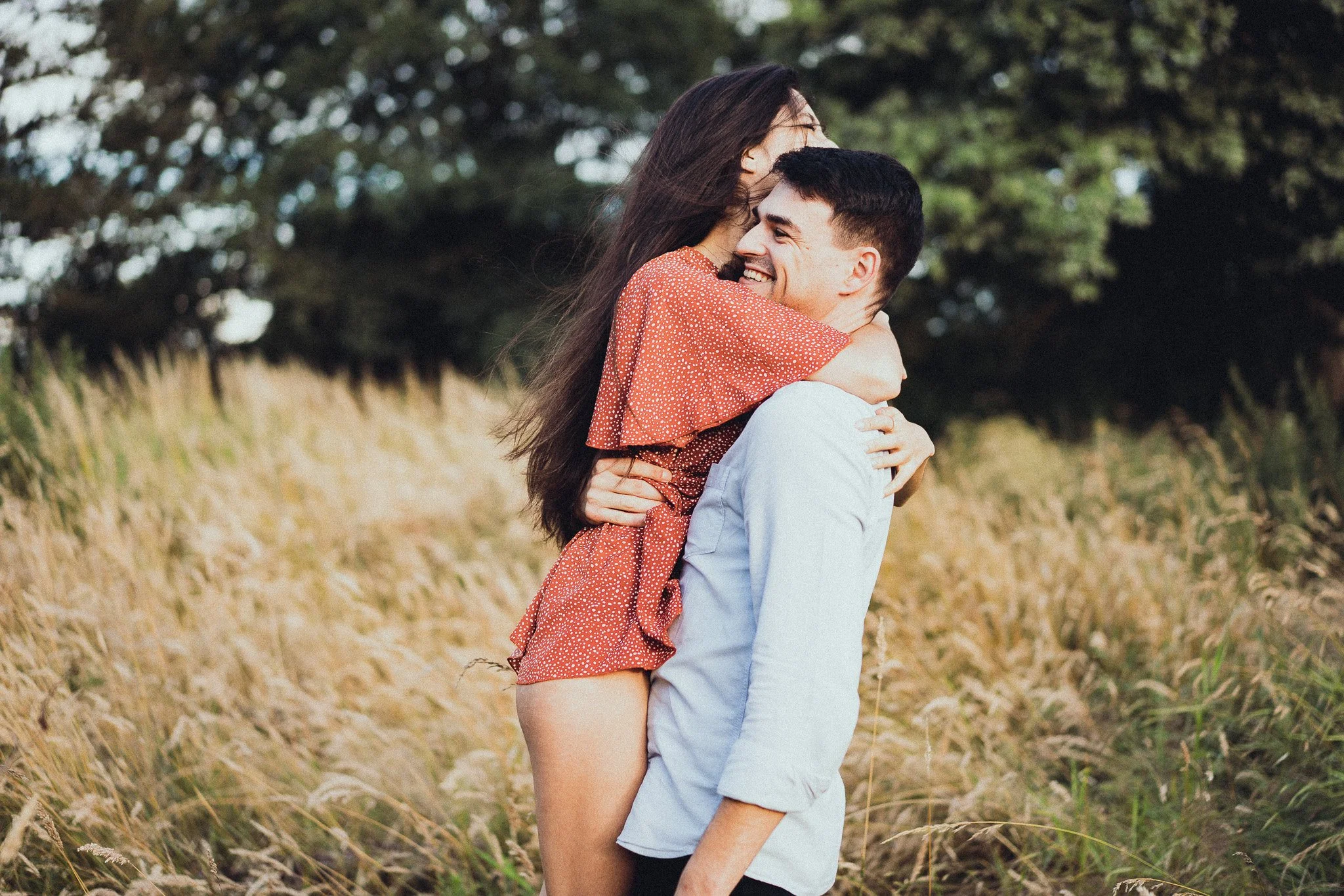Un couple souriant en plein air, l'homme portant une chemise blanche et la femme en robe rouge à pois, en train de s'étreindre dans un champ avec des arbres en arrière-plan.