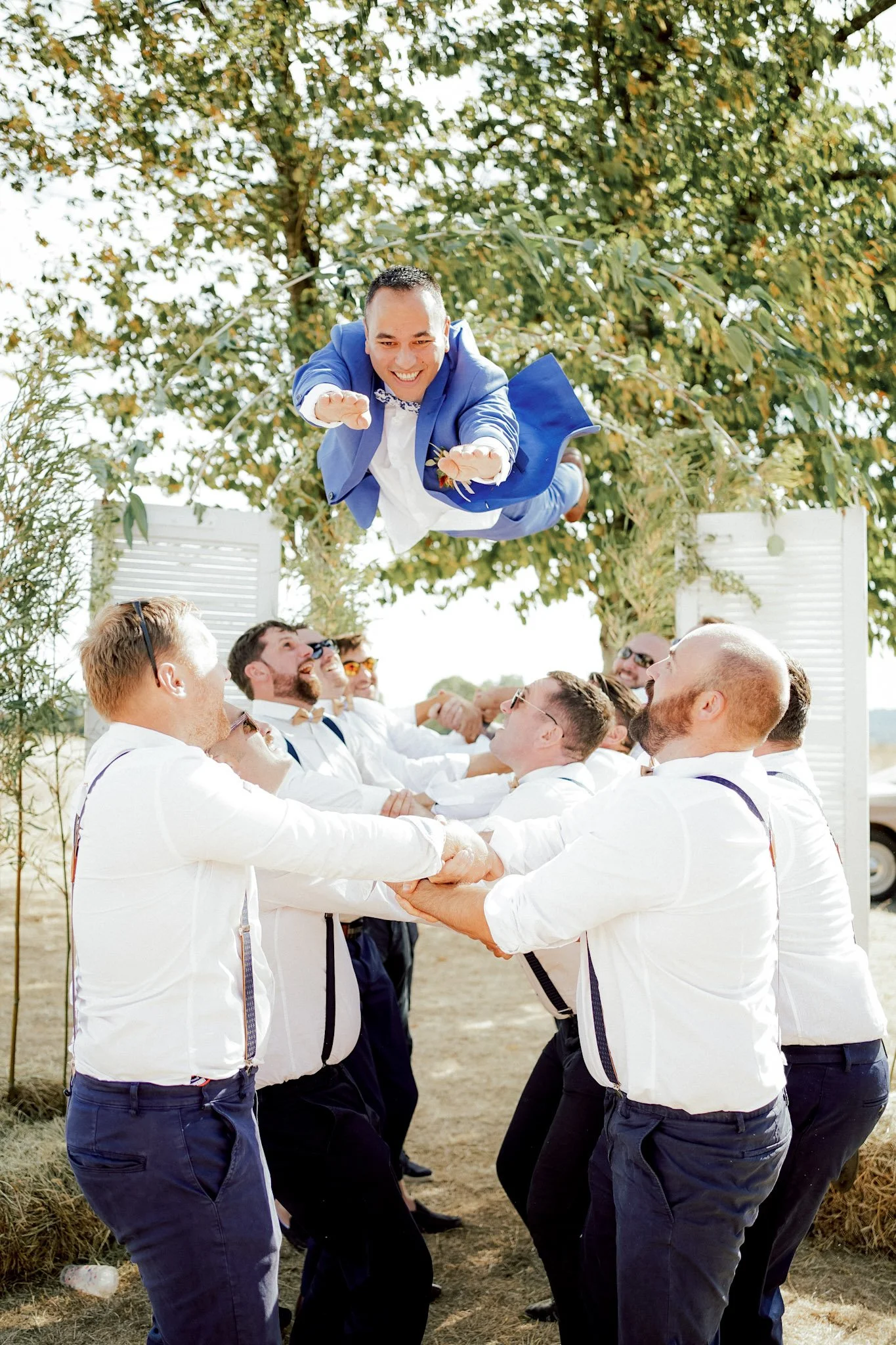 Un groupe d'hommes habillés en blanc et en chemises noirs font une danse de mariage en tenant un homme en costume bleu qui saute à travers leurs bras, sous un ciel clair avec des arbres en arrière-plan.