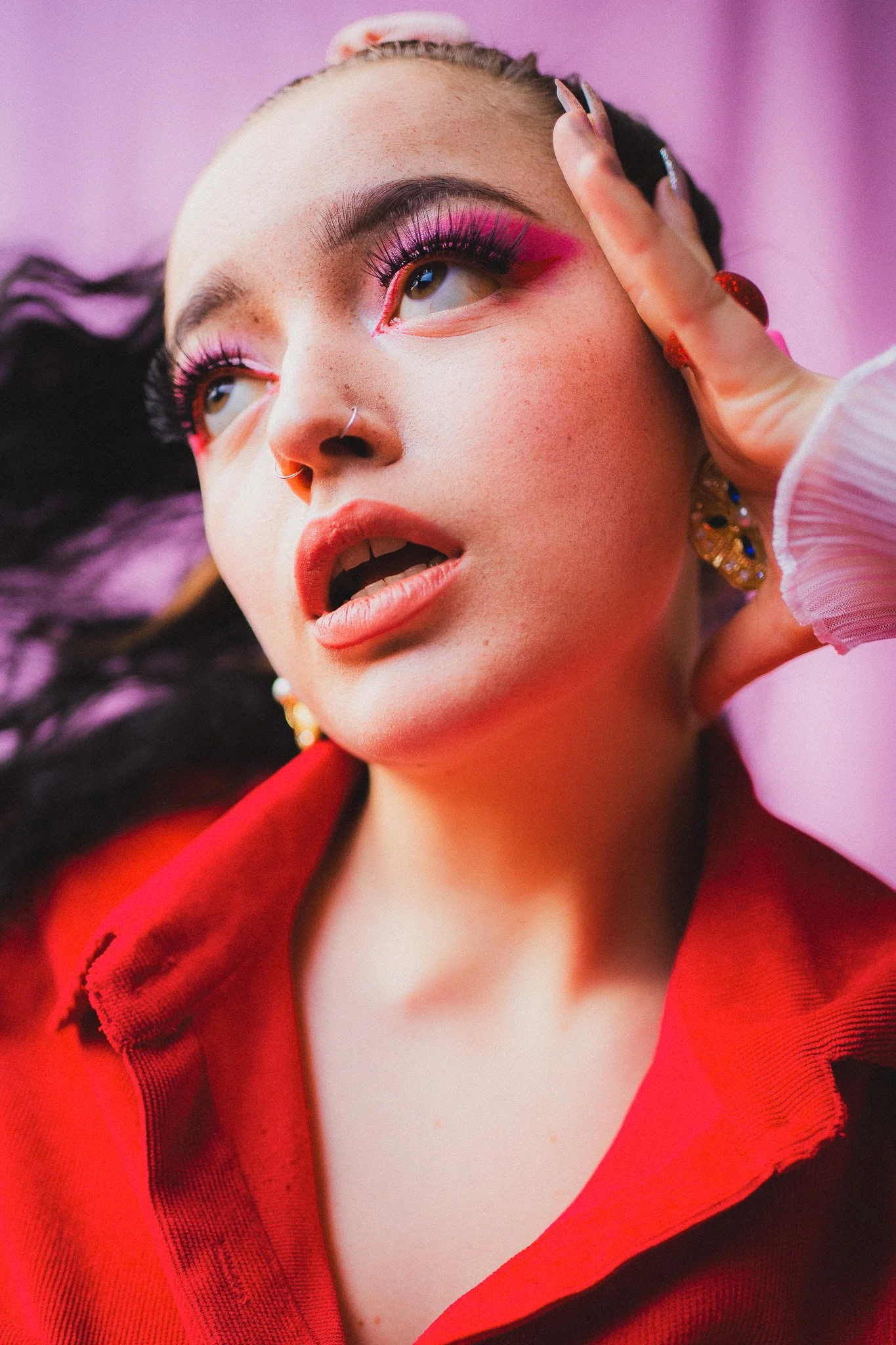 Jeune femme avec maquillage vif, portant une veste rouge, jouant avec ses cheveux devant un fond rose.