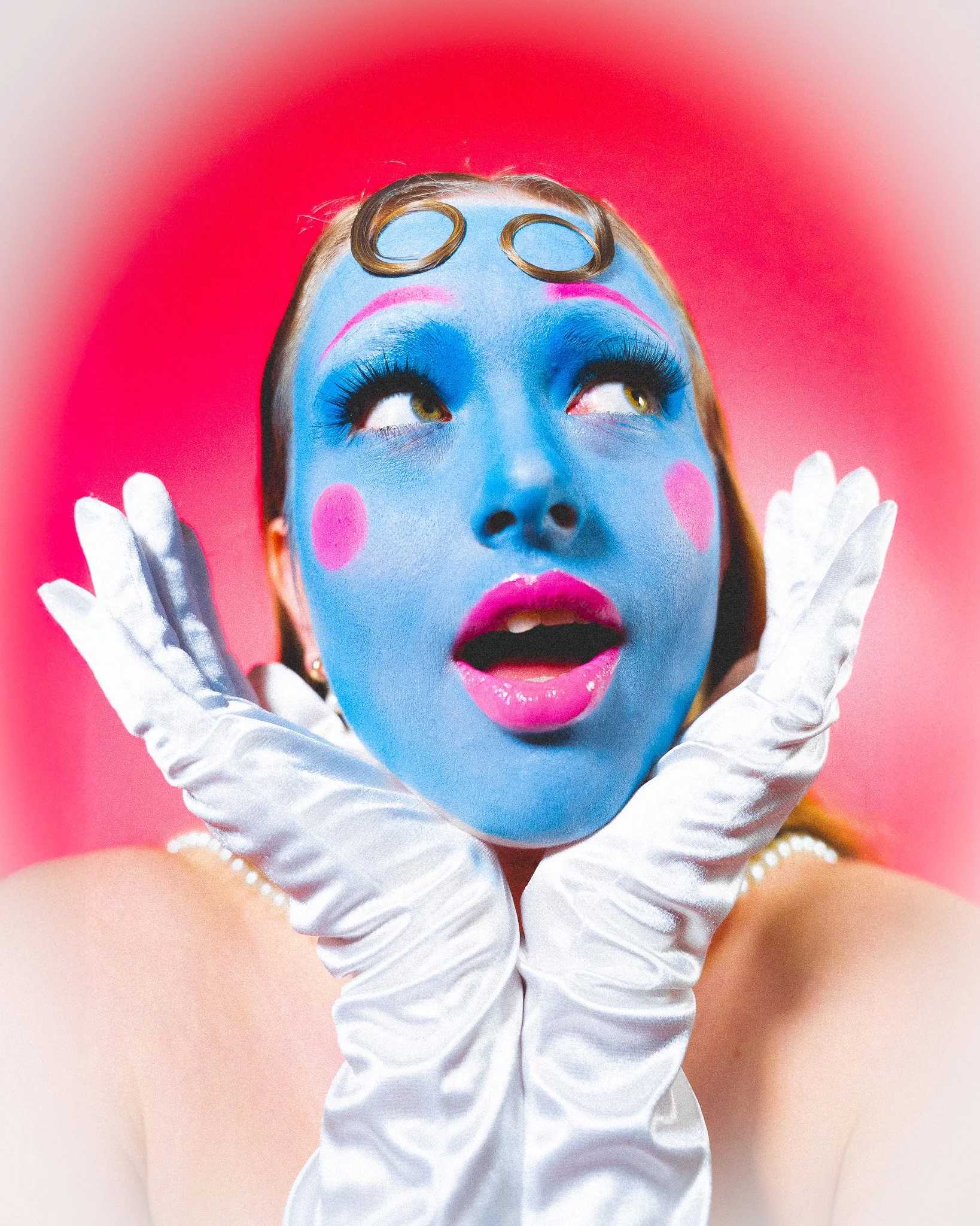 Jeune femme avec un visage peint en bleu, maquillage coloré, portant des gants de soie blancs, regardant vers le haut, avec un fond rose.