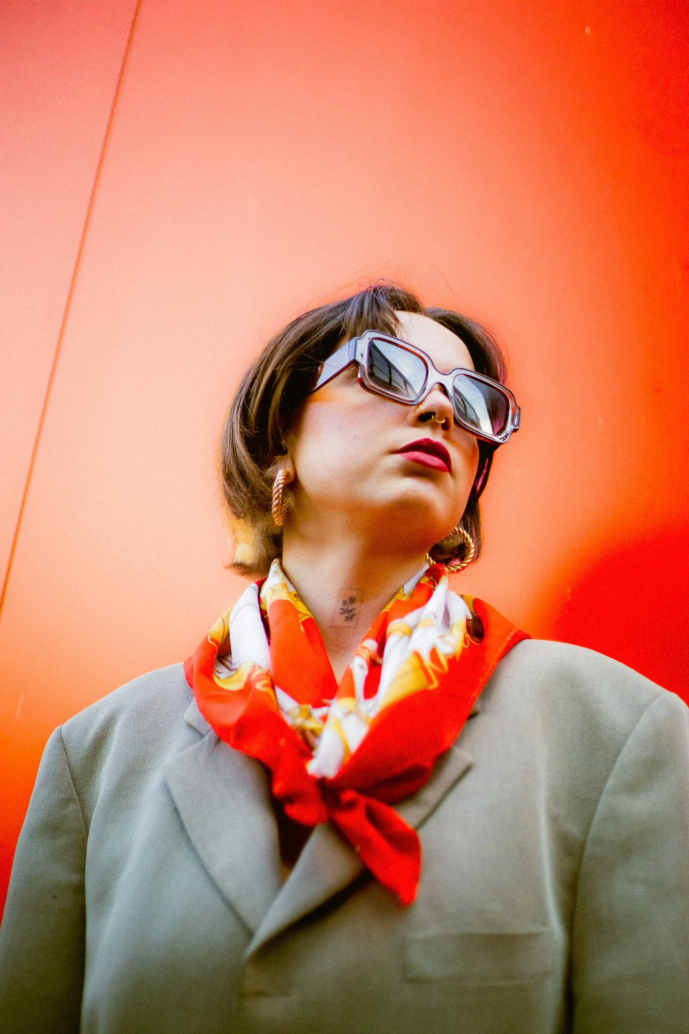 Une femme avec des lunettes de soleil, des boucles d'oreilles criantes, un foulard coloré, vêtue d'un trench beige, se tenant devant un mur orange.