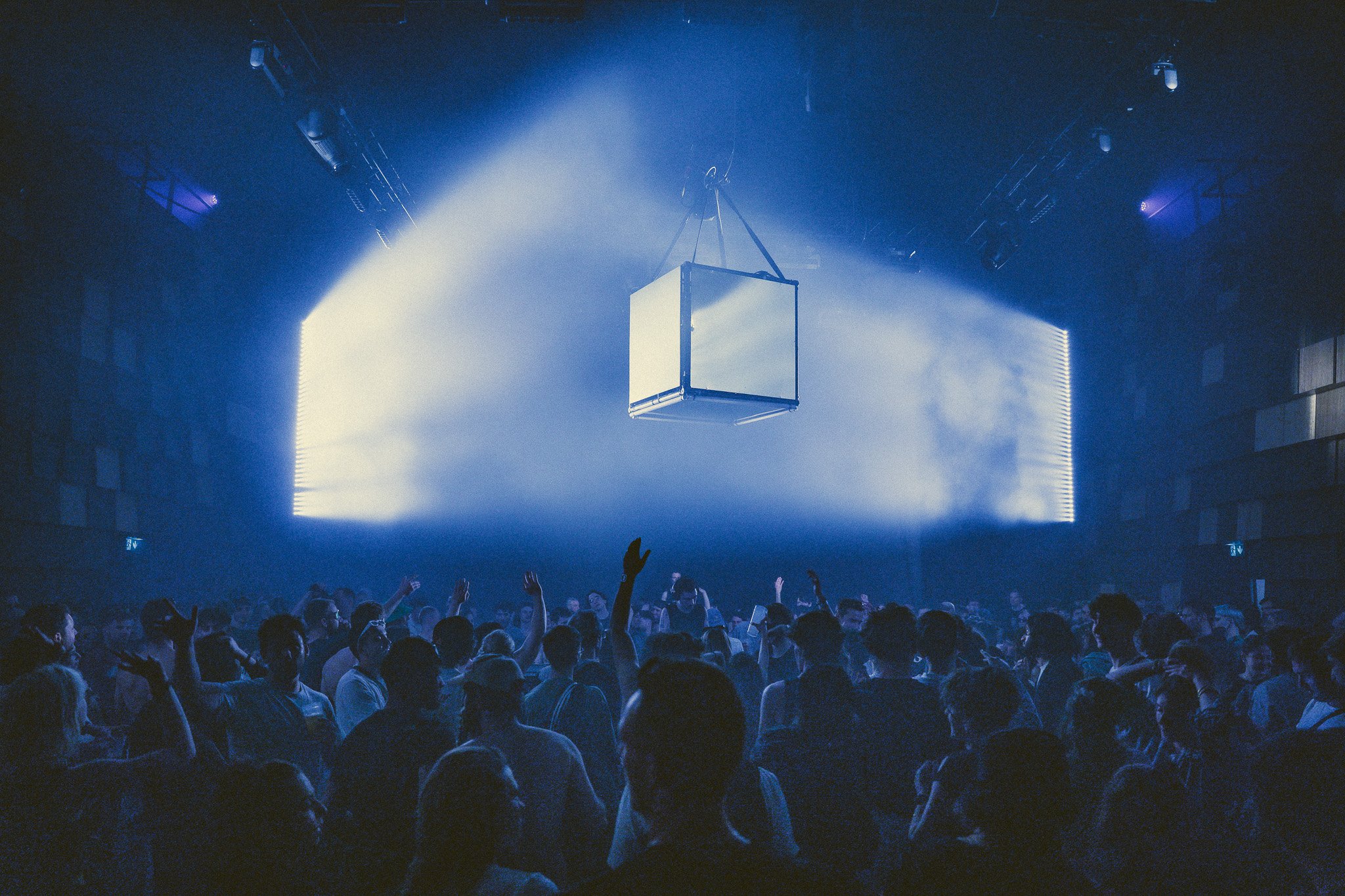 Une foule de personnes danse dans une discothèque ou un club, sous une lumière bleue avec des effets lumineux, avec un grand cube suspendu au plafond.