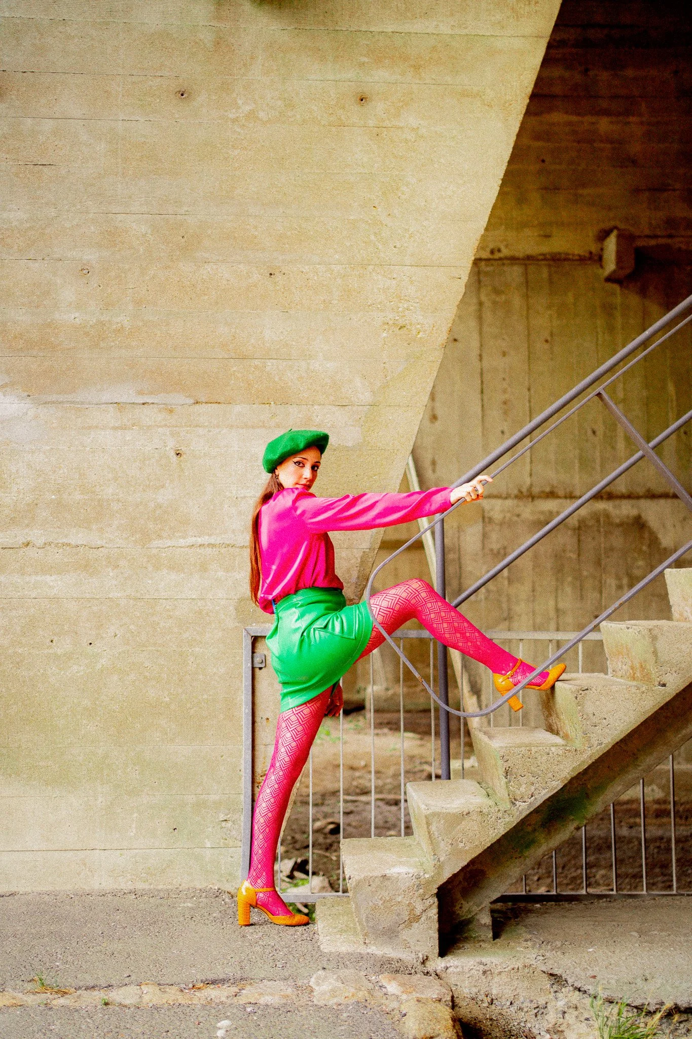 Une femme en tenue colorée grimpe les escaliers en béton, portant une chemise rose, une jupe verte, des collants roses avec motif, des talons oranges, et un béret vert dans un environnement urbain industriel.