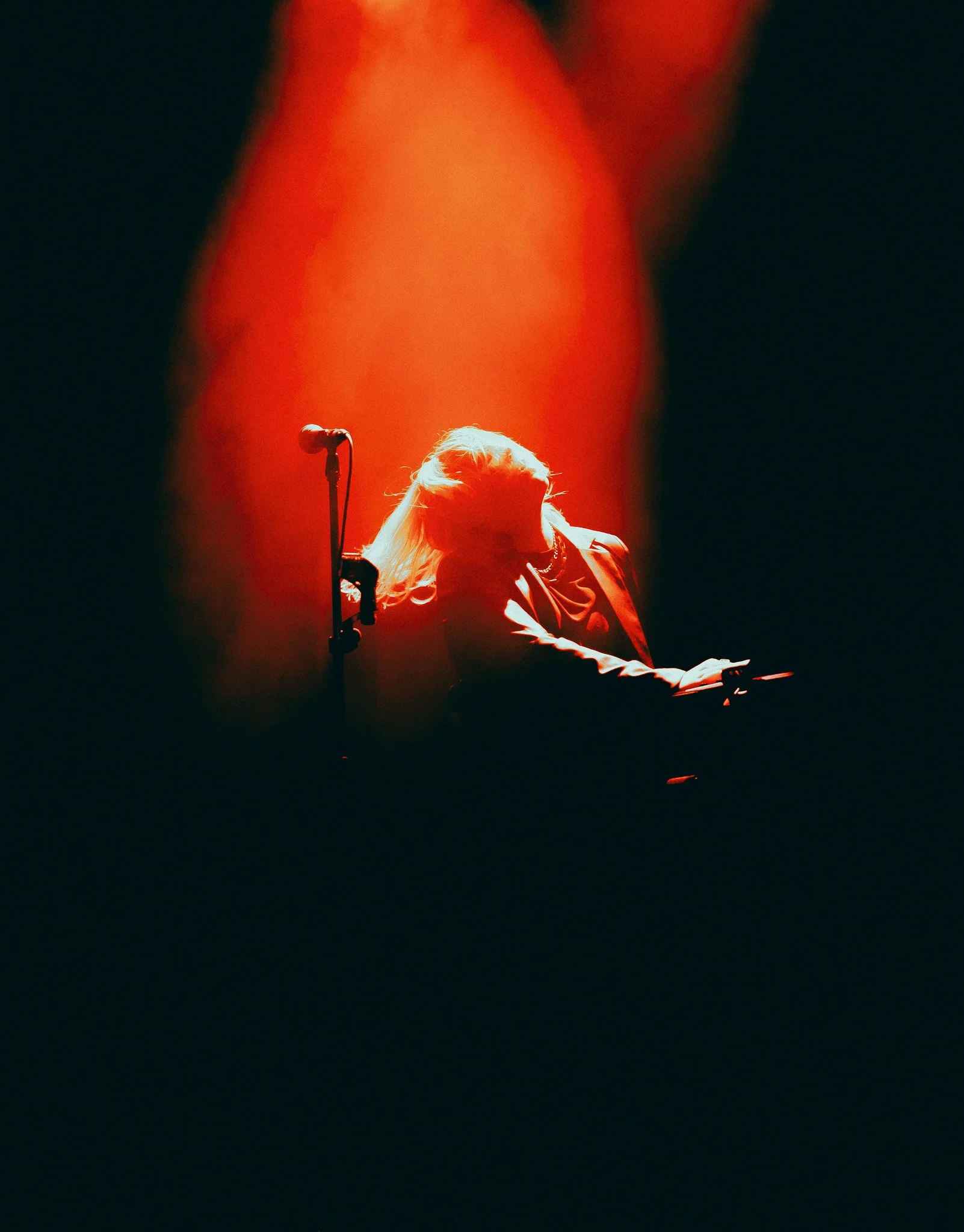 Une personne avec des cheveux blonds jouant du clavier sur scène, éclairée par un éclairage rouge, avec un microphone à proximité, dans un environnement sombre et dramatique.