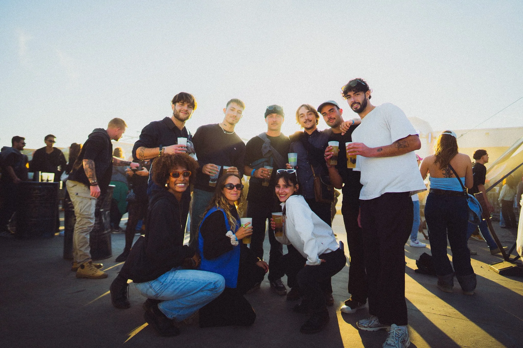 Groupe de jeunes amis souriants posant pour une photo lors d'un événement en plein air, certains tenant des boissons. Le soleil couchant éclaire la scène, dans une ambiance festive.