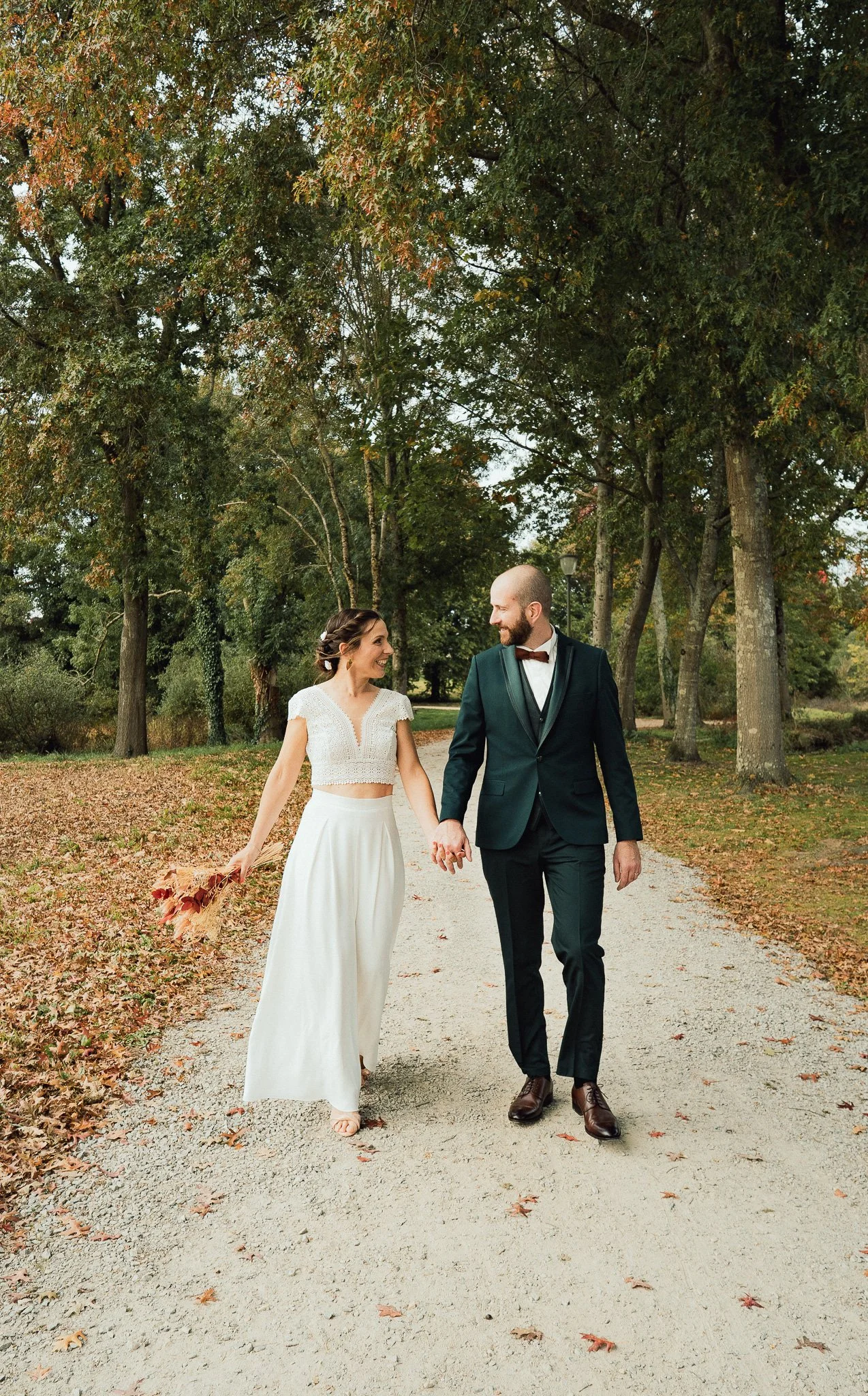 Un couple habillé en mariage marche dans un parc entouré d'arbres en automne, se tenant la main et se regardant avec sourire.