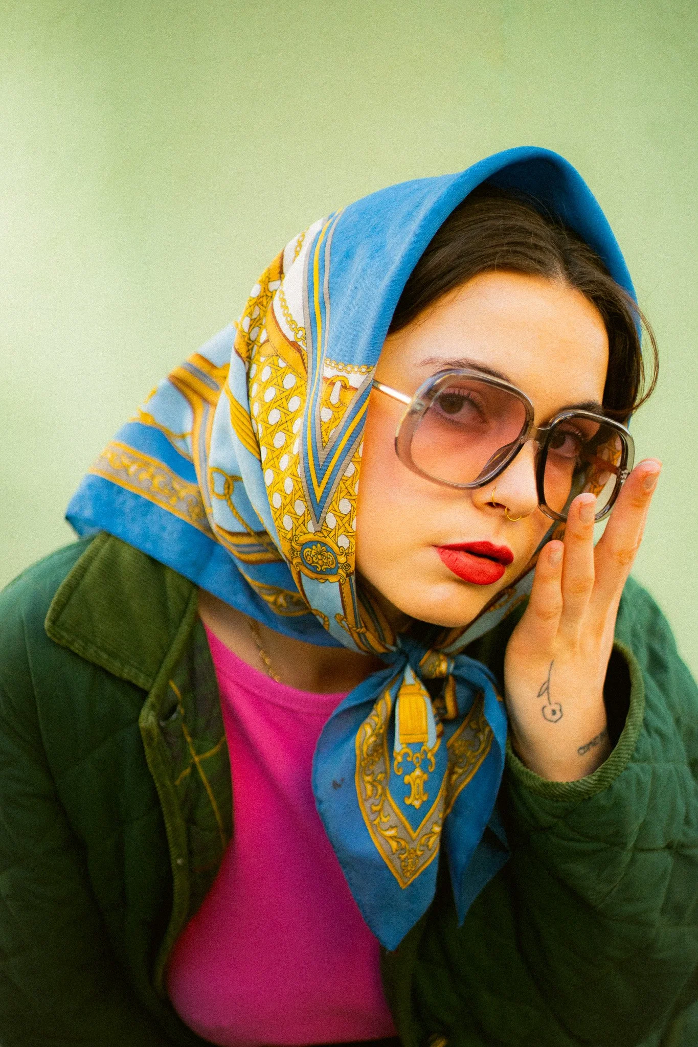 Jeune femme avec un foulard coloré sur la tête portant des lunettes et un maquillage rouge, touchant son visage avec une main tatouée.