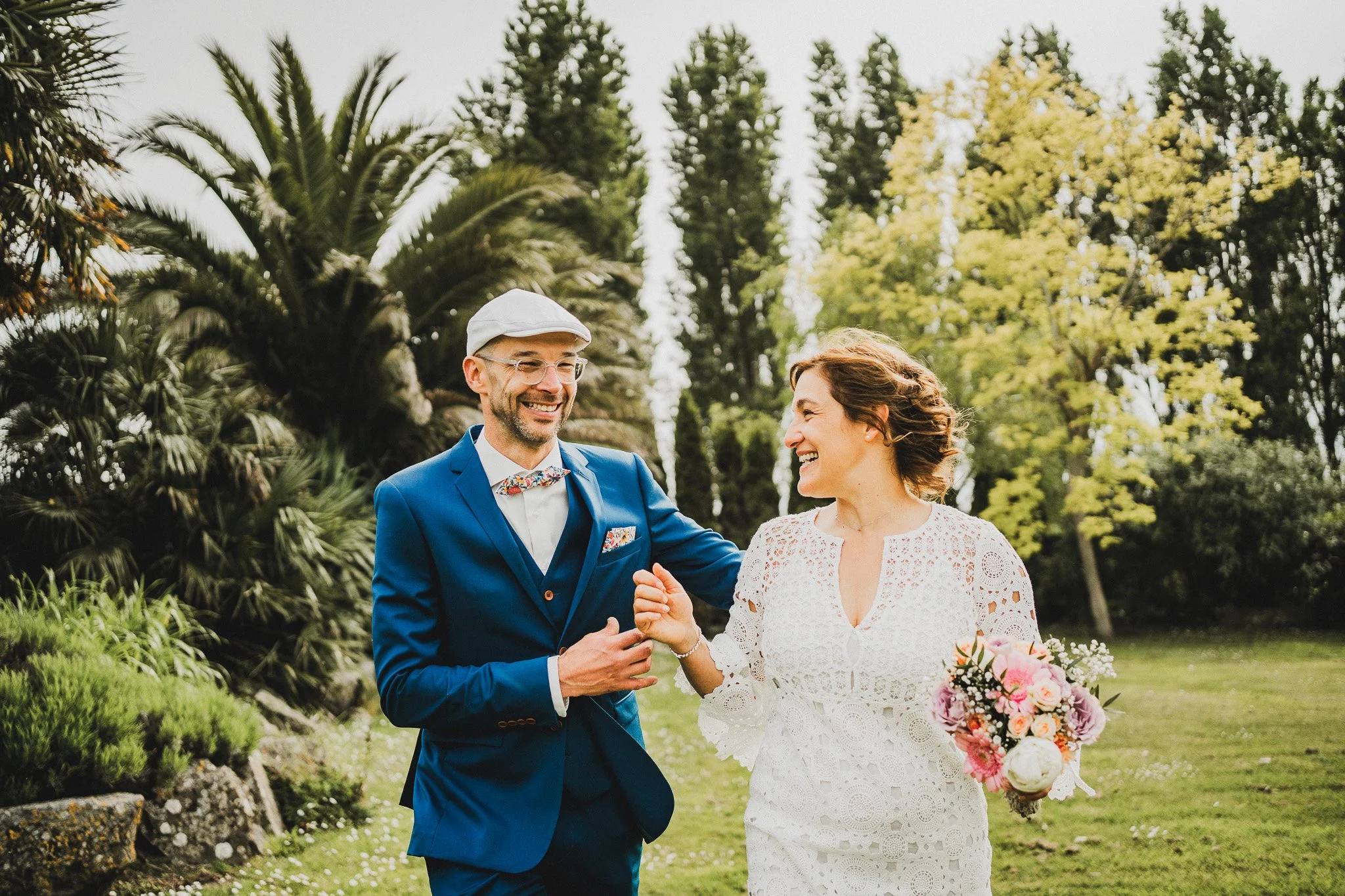 Un couple de mariage souriant dans un jardin, l'homme en costume bleu et la femme en robe blanche avec un bouquet de fleurs.
