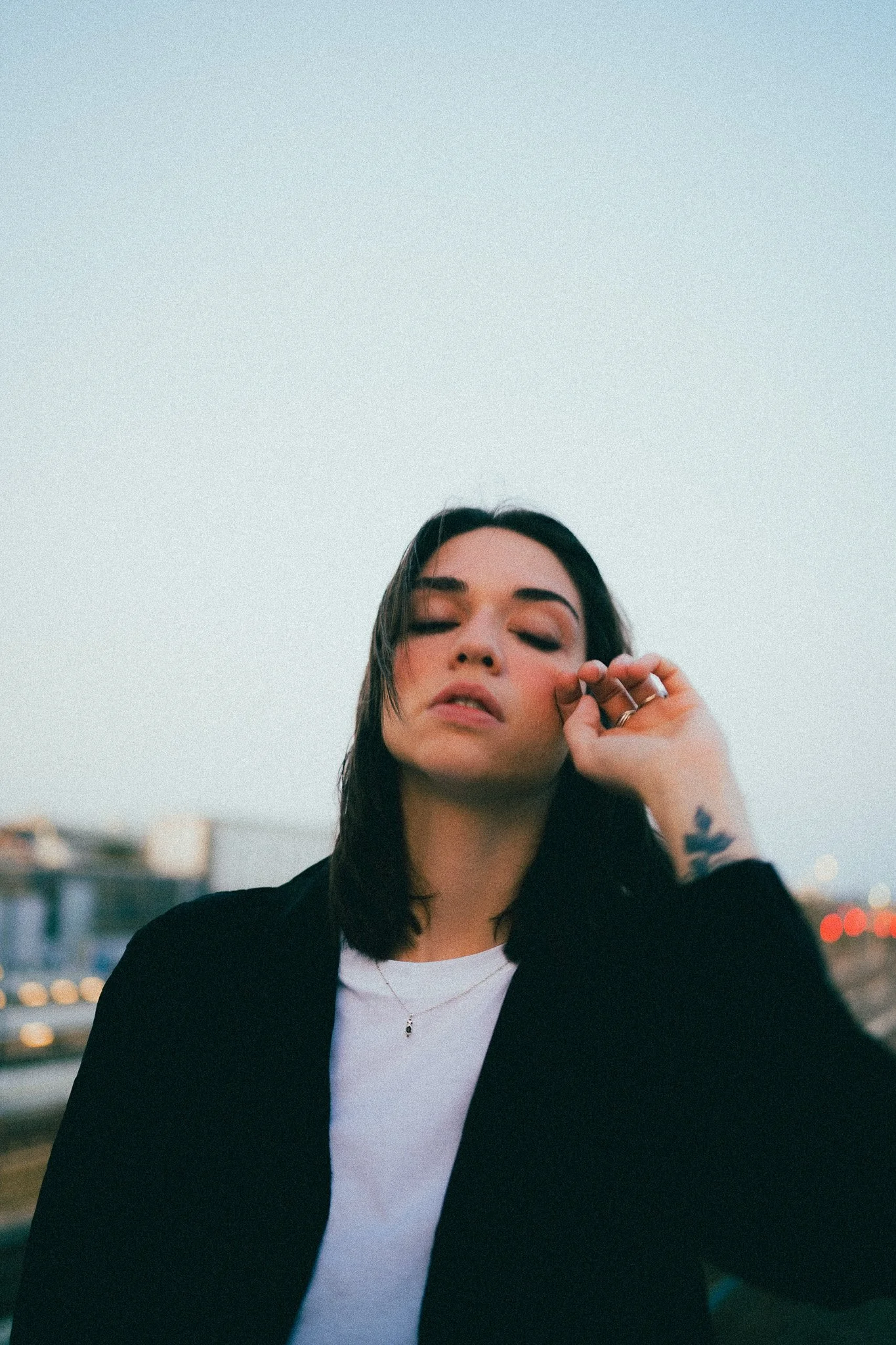 Jeune femme aux cheveux noirs, portant un haut blanc et un blazer noir, avec un tatouage sur le bras, en extérieur, à la tombée de la nuit.