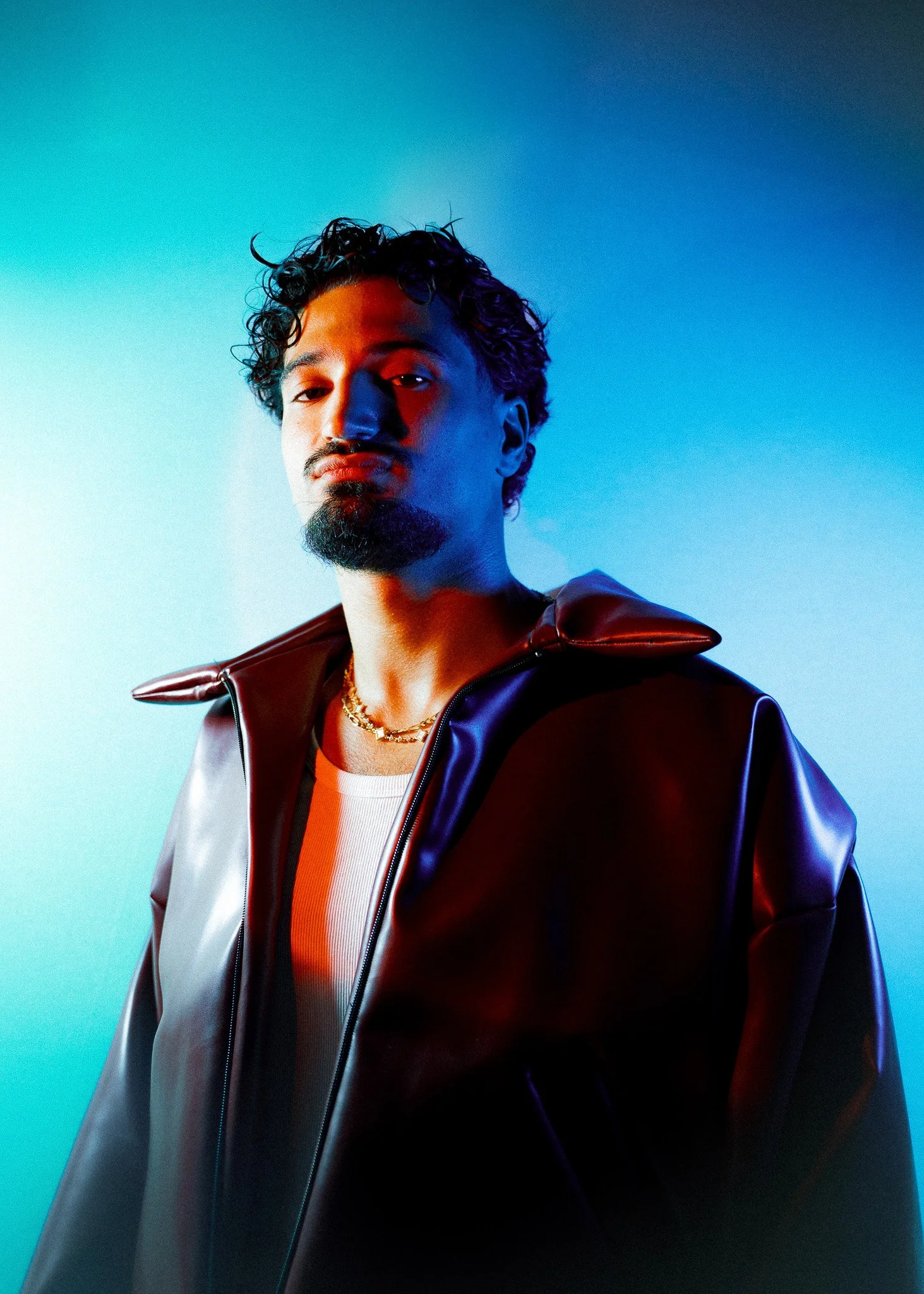 Jeune homme avec barbe, portant une veste en cuir noir avec des épaules épaisses, un t-shirt blanc, et une chaîne en or, éclairé par une lumière bleue et rouge dans un fond de couleur unie.