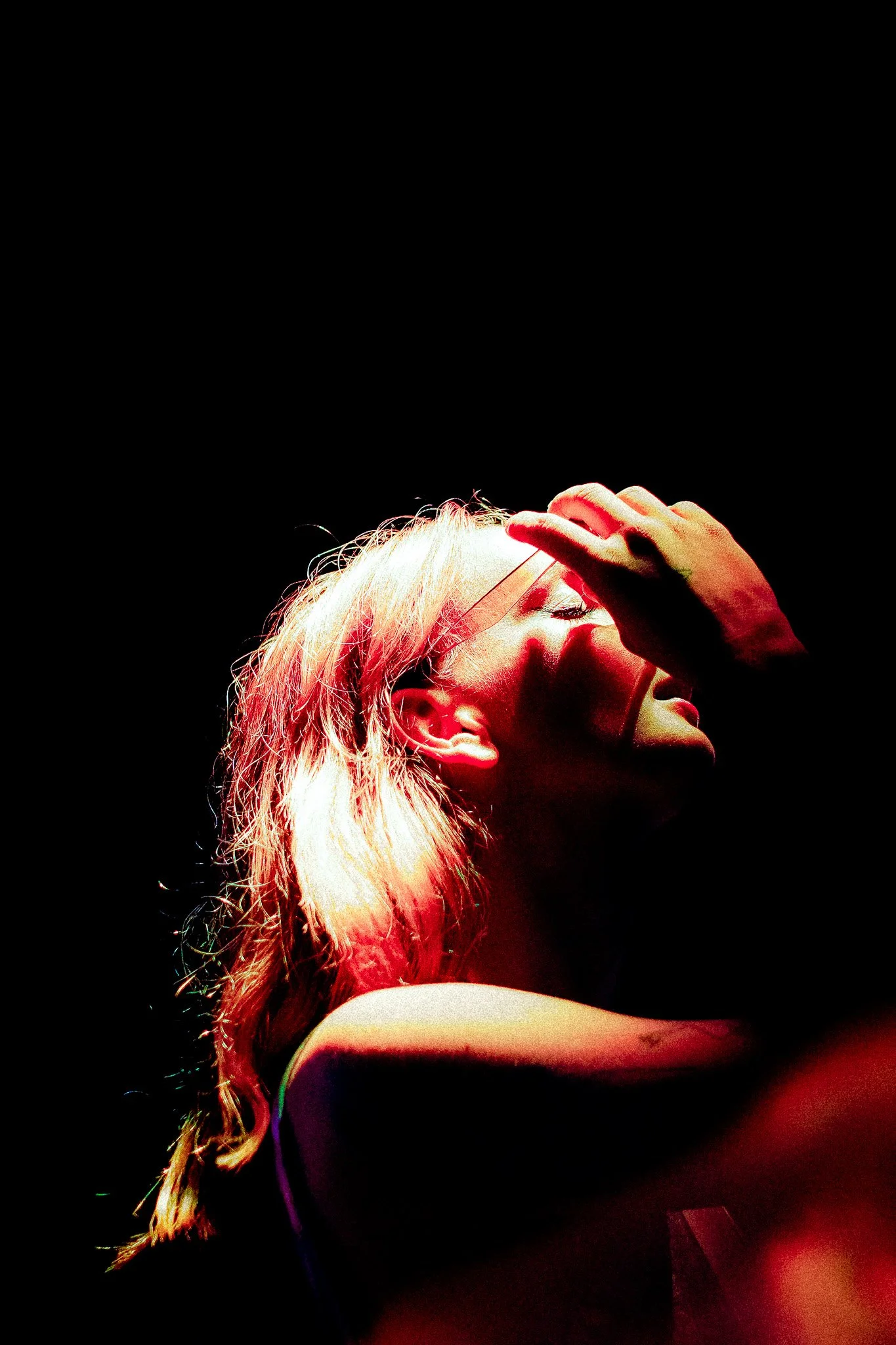 Une femme blonde avec des cheveux ondulés, portant des lunettes, la main sur le front, l'air pensif ou stressé, dans un éclairage rouge sur fond noir.