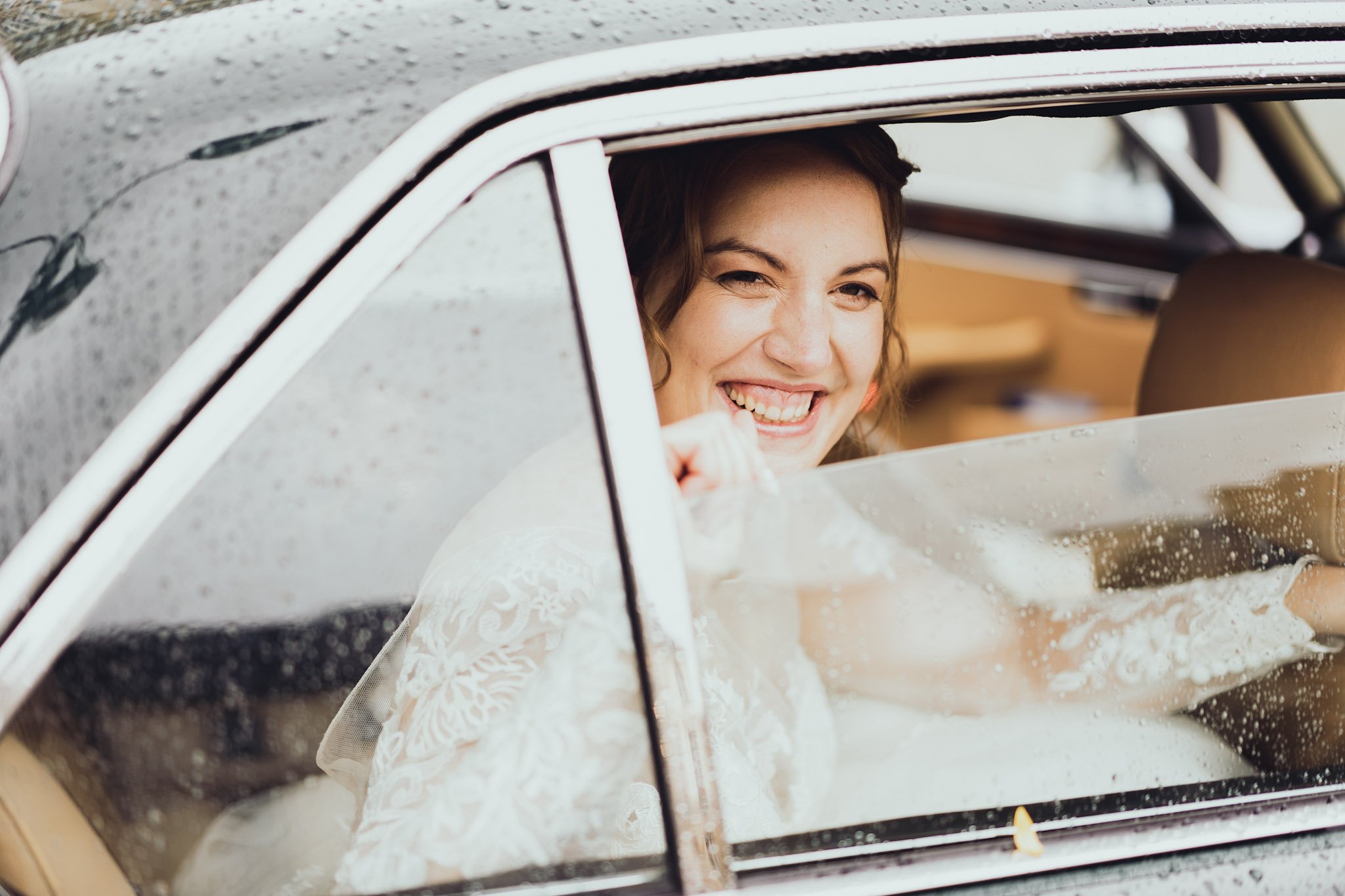 Une femme souriante, probablement une mariée, assise dans une voiture chaude et humide, regardant par la fenêtre.
