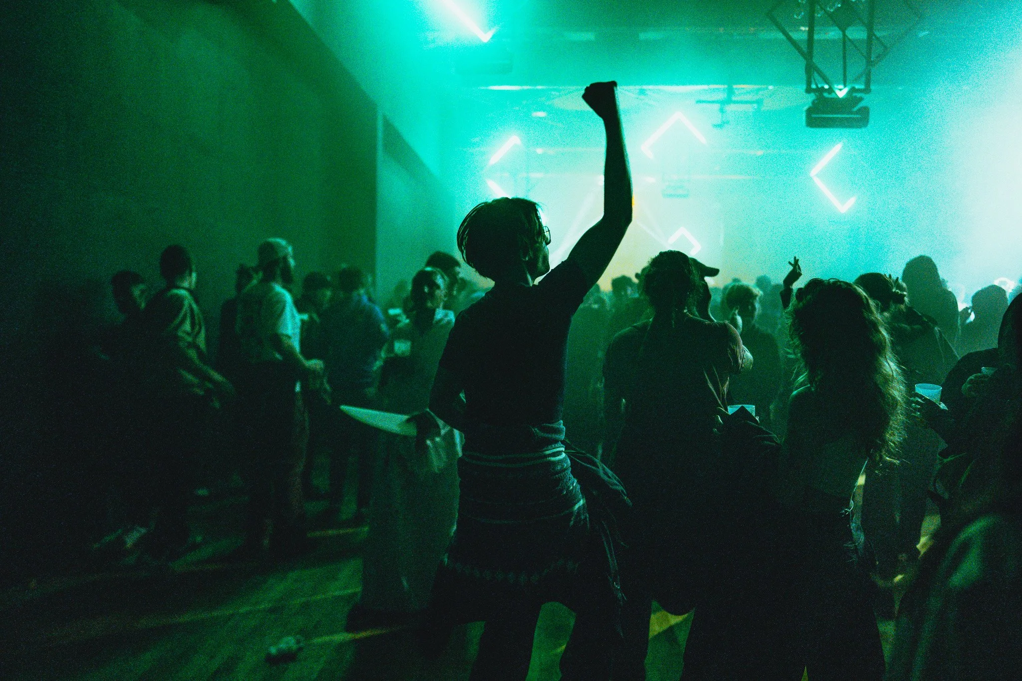 Une scène de fête ou de concert avec des personnes dansant et socialisant dans une ambiance sombre illuminée par des lumières de couleur verte et des néons. Un homme au centre lève le bras, tenant une assiette, entouré d'autres personnes.