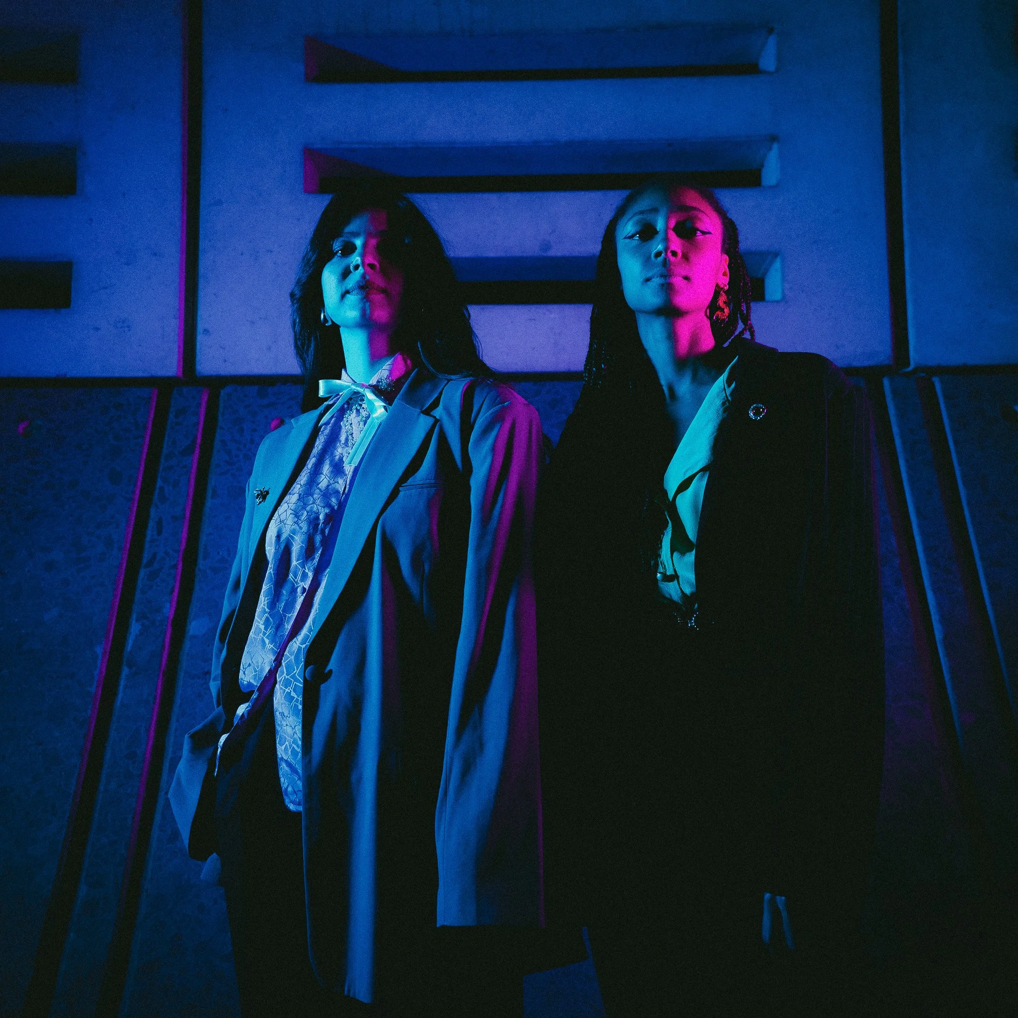 Deux femmes debout dans une pièce sombre, éclairées par des lumières roses et bleues, portant des vêtements élégants.