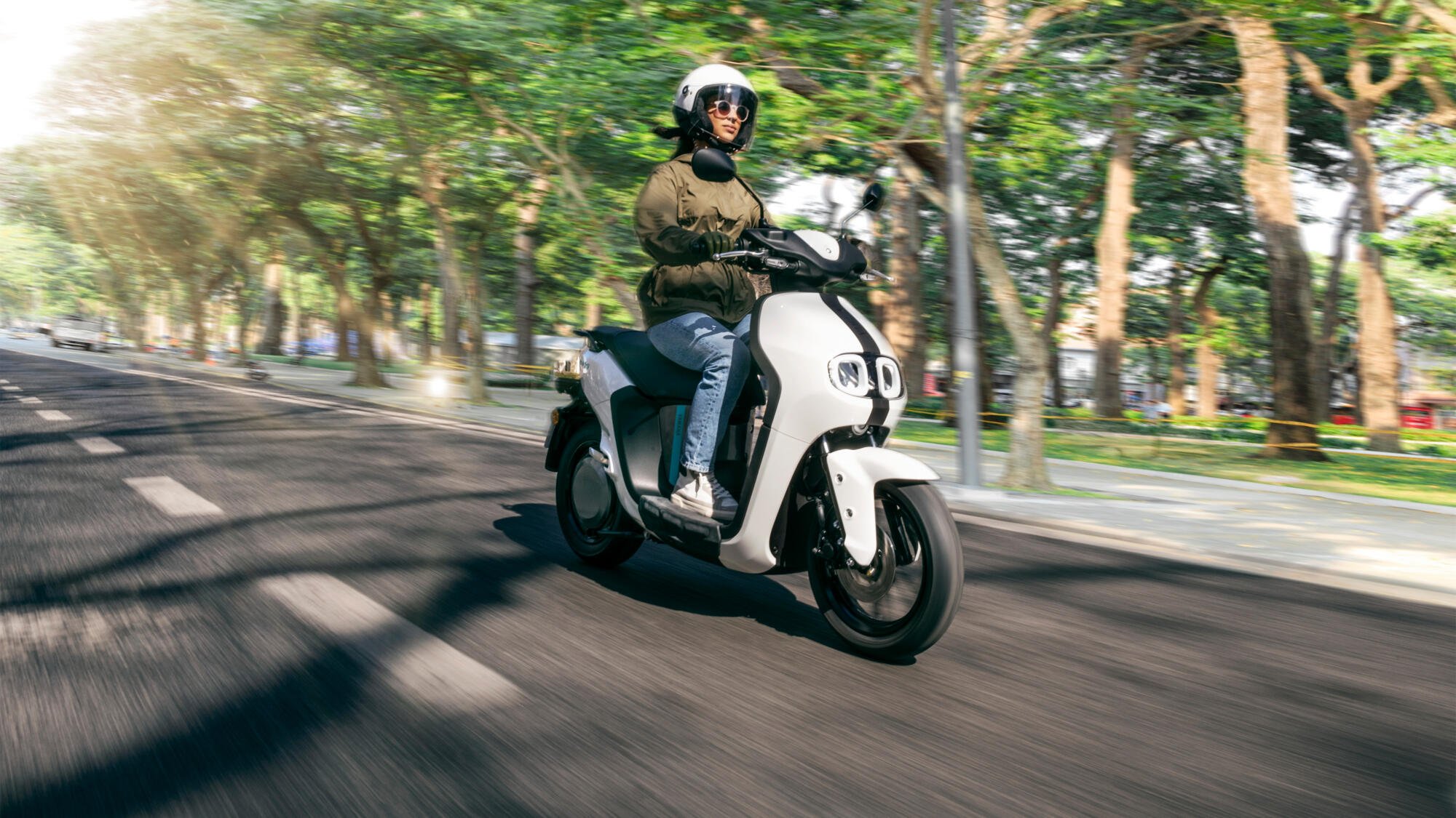 Une femme portant un casque et des lunettes de soleil, chevauchant une trottinette électrique blanche en mouvement sur une route entourée d'arbres.