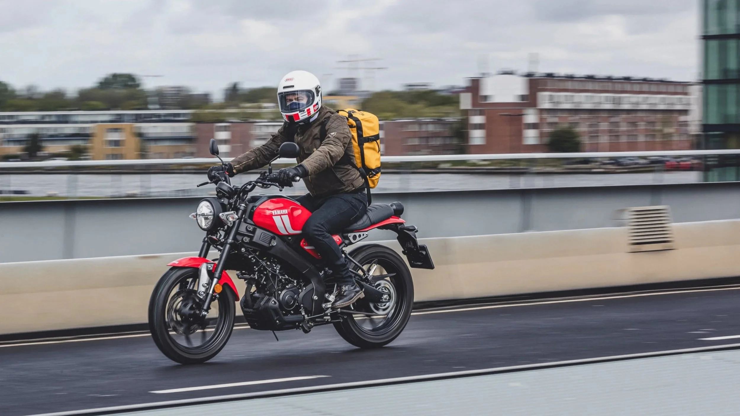 Un homme en casque blanc et veste marron roulant à moto rouge sur un pont urbain.