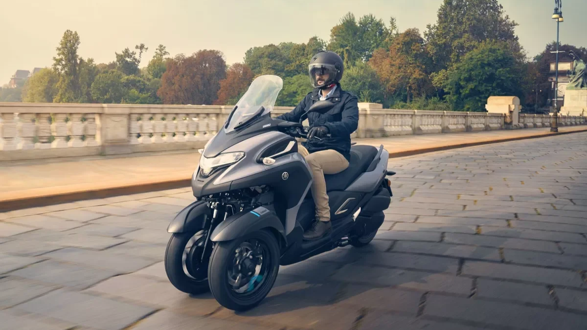 Une personne portant un casque de moto et une veste noire, chevauchant un scooter noir sur un pont en pierre avec un décor urbain et des arbres en arrière-plan.