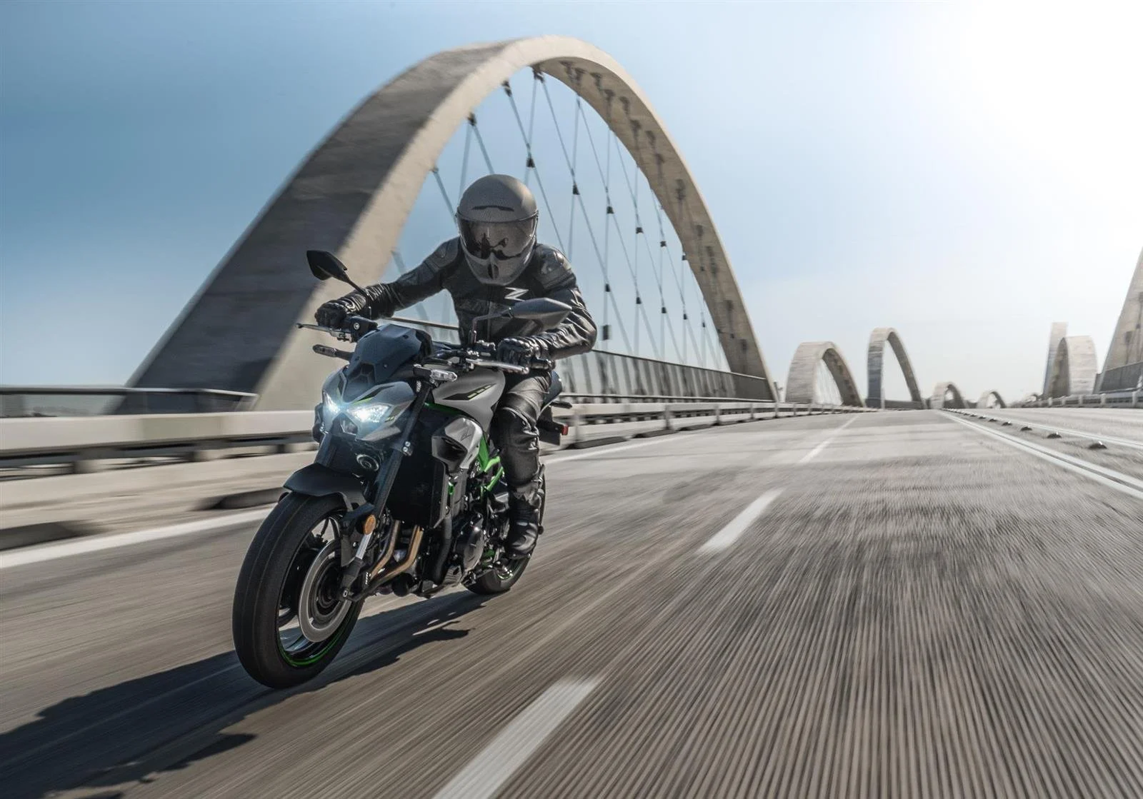 Un motard en noir roulant sur une route traversant un pont moderne en arc, sous un ciel clair.