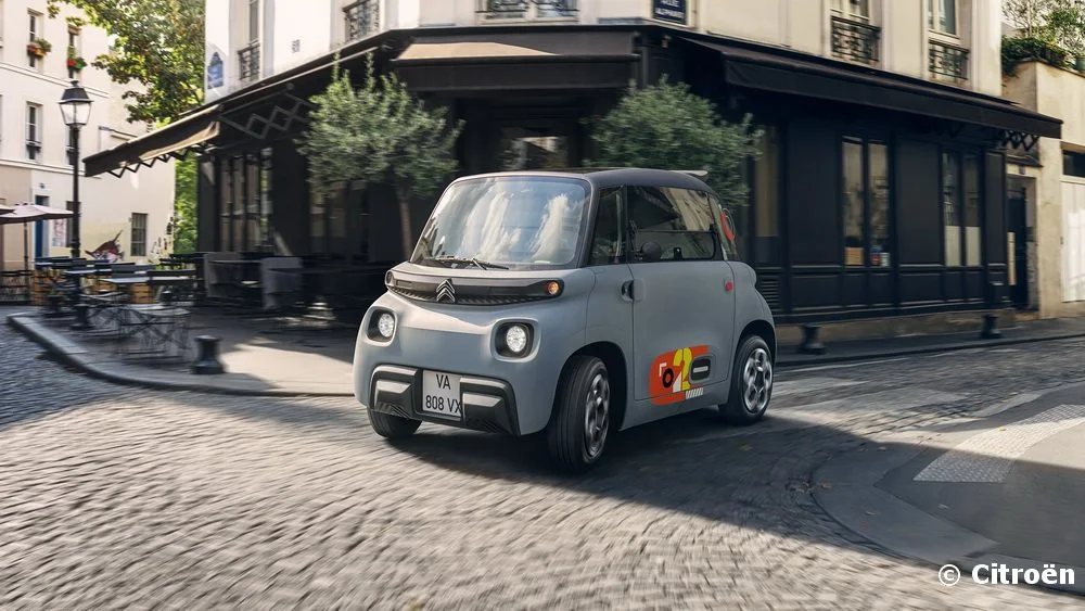 Voiture électrique compacte de Citroën, couleurs gris clair avec graphique coloré sur la porte, en mouvement dans une rue urbaine pavée, bâtiments anciens et terrasses de café en arrière-plan.