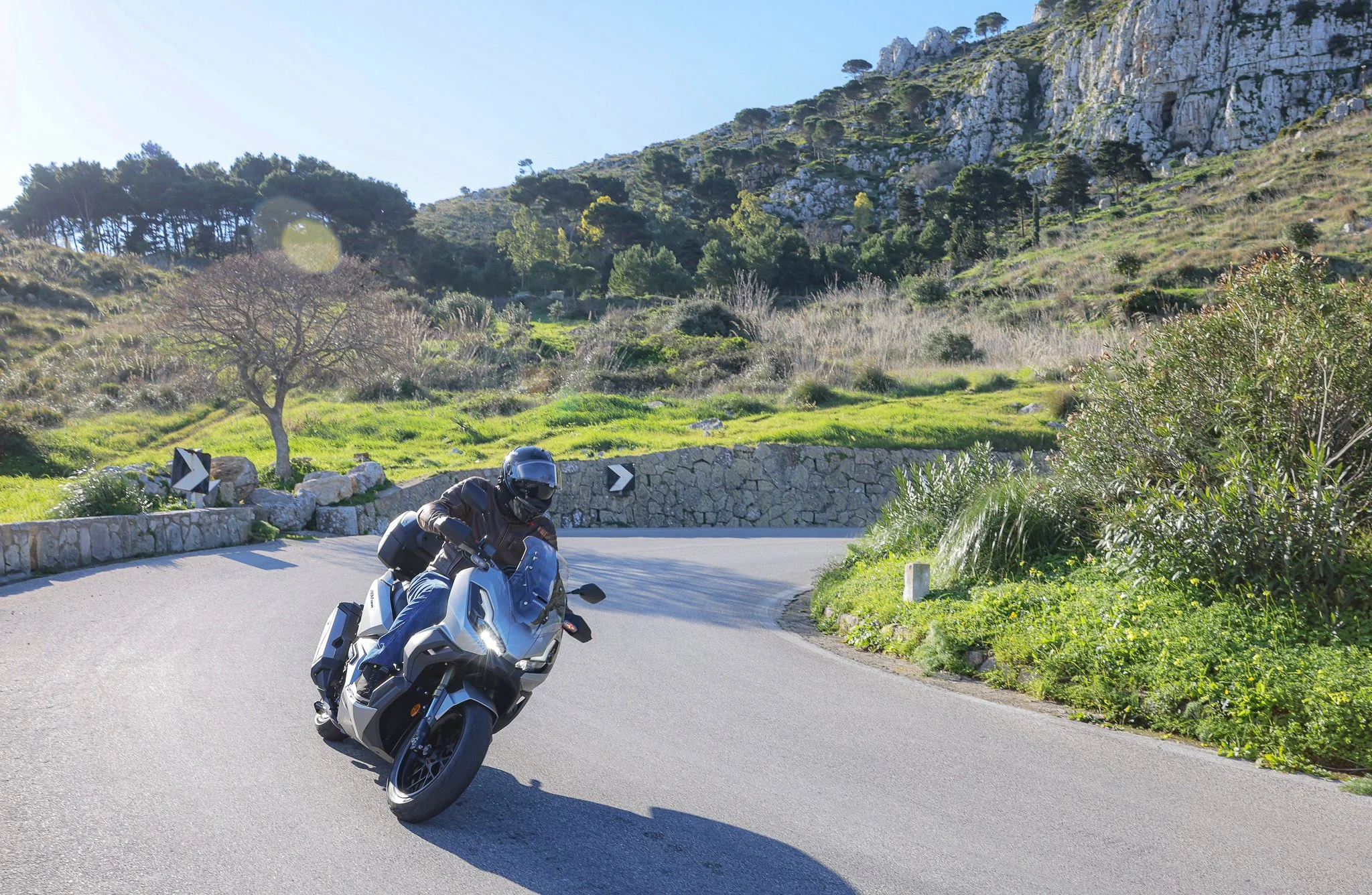 Un motard portant un casque noir et une veste en cuir, chevaucheant une moto blanche sur une route sinueuse entourée de verdure et de montagnes en arrière-plan.