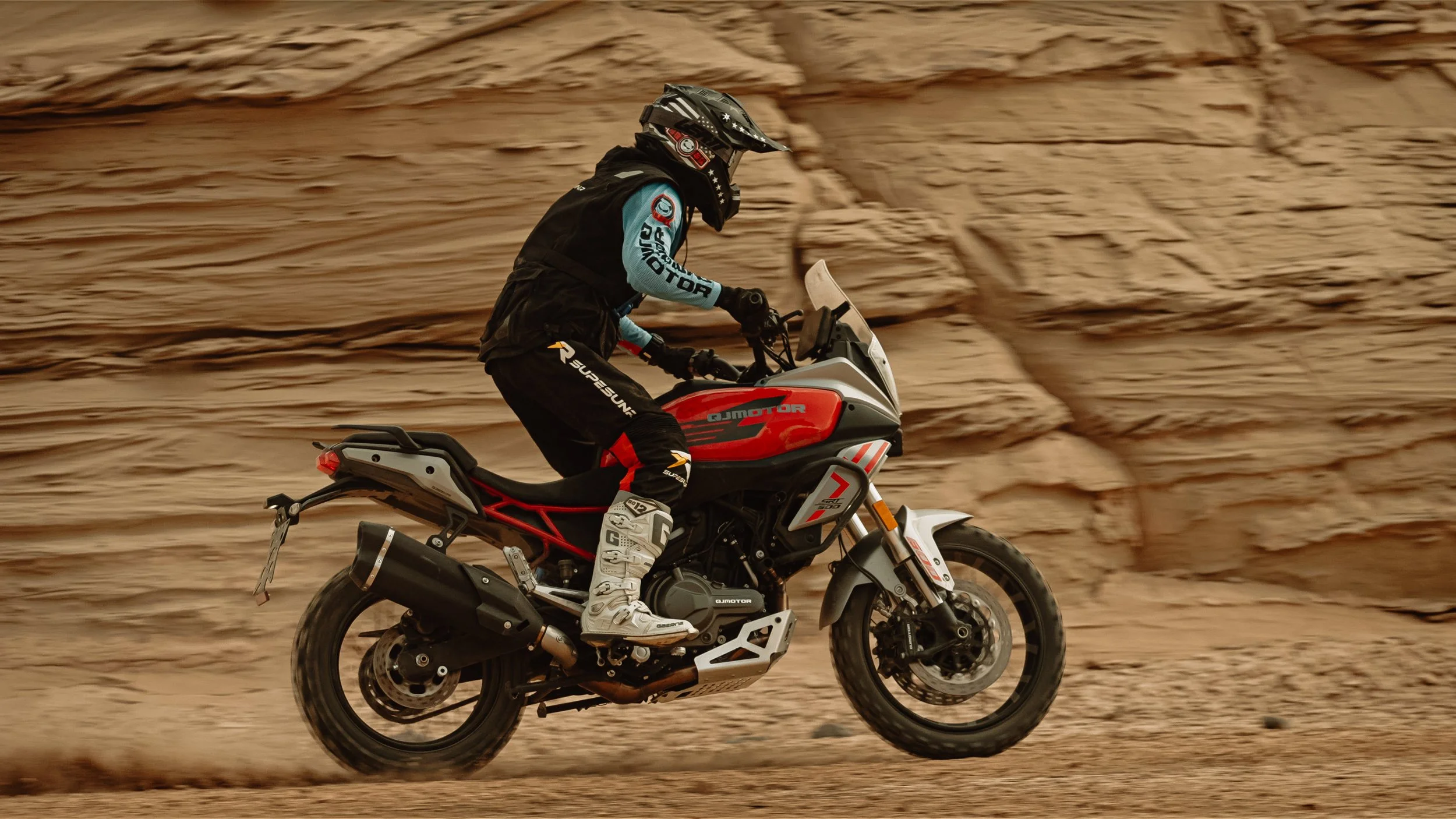Une personne en équipement de moto tout-terrain, portant un casque, des gants, une veste et des bottes, roulant sur une moto dans un environnement désertique rocheux.