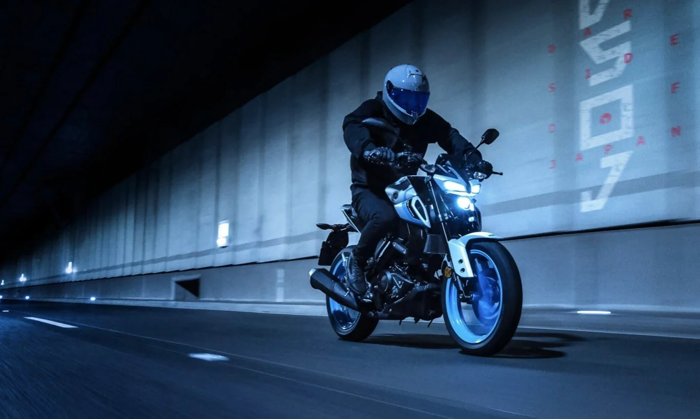 Un motocycliste portant un casque et un équipement noir, roulant dans un tunnel sombres avec des murs en béton et des inscriptions lumineuses sur la paroi, la moto étant éclairée et moderne.