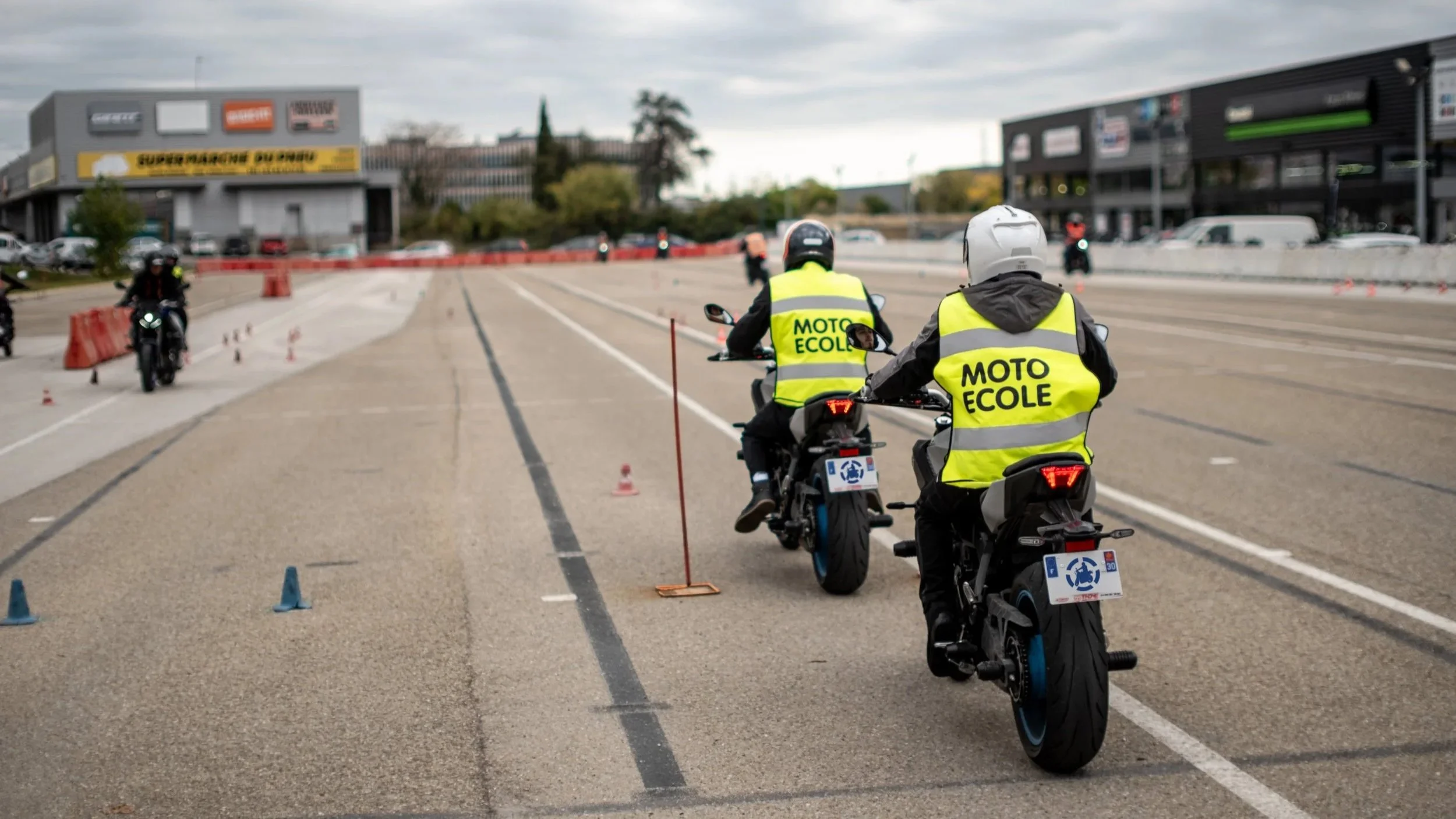 Deux motards portant des vestes jaunes avec l'inscription 'MOTO ÉCOLE' participent à une formation sur un grand espace pavé, avec quelques cônes de signalisation et d'autres motos en arrière-plan.