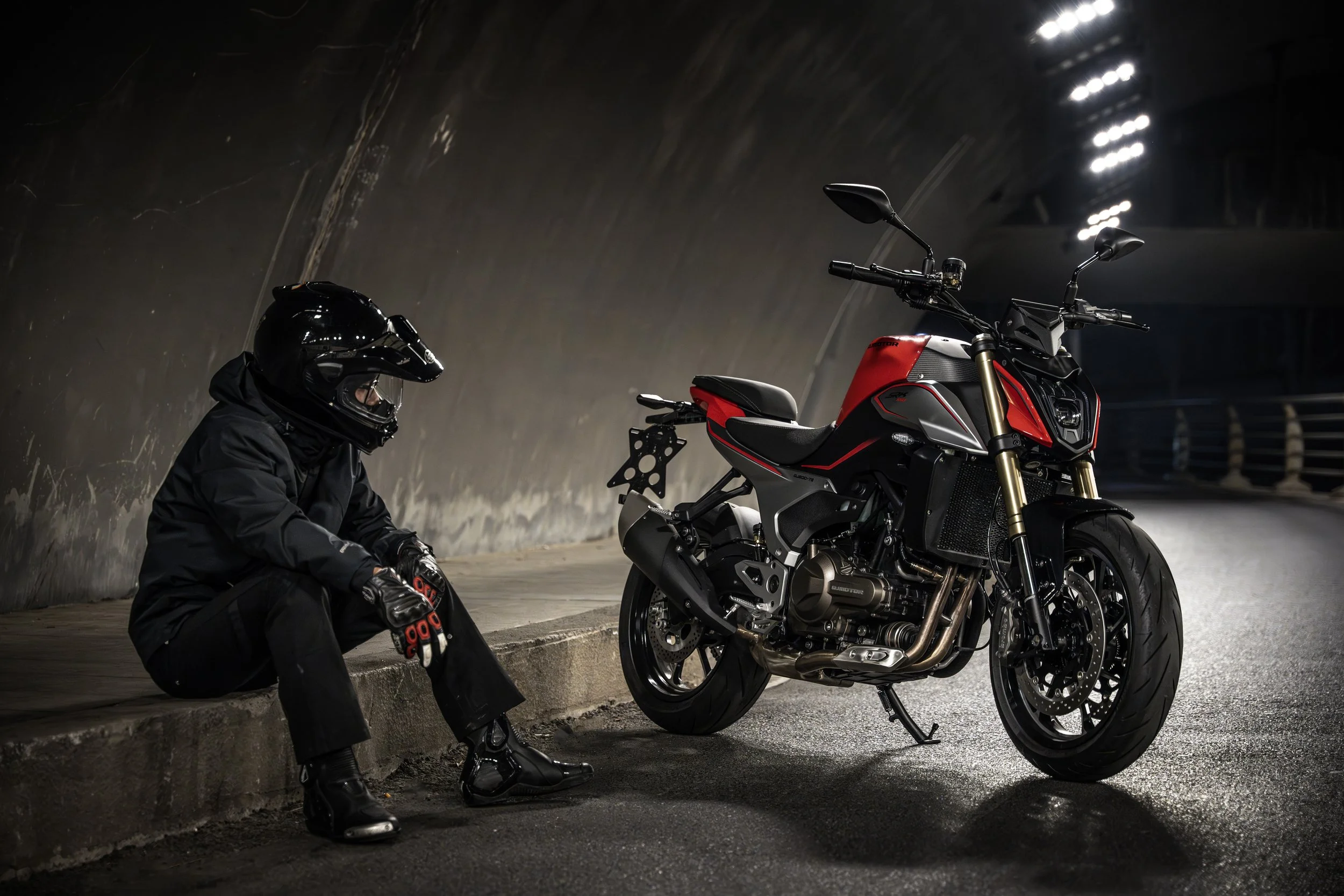 Une personne en équipement de moto, assise sur le sol d'un tunnel, près d'une moto noire et rouge.