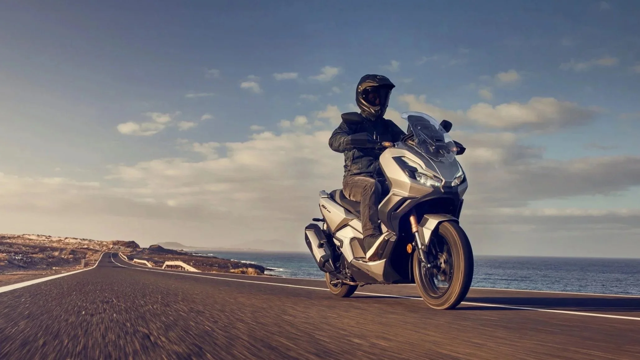 Motard roulant sur une route côtière au coucher du soleil, vue du bas de la moto, avec mer et ciel dégagé en arrière-plan.