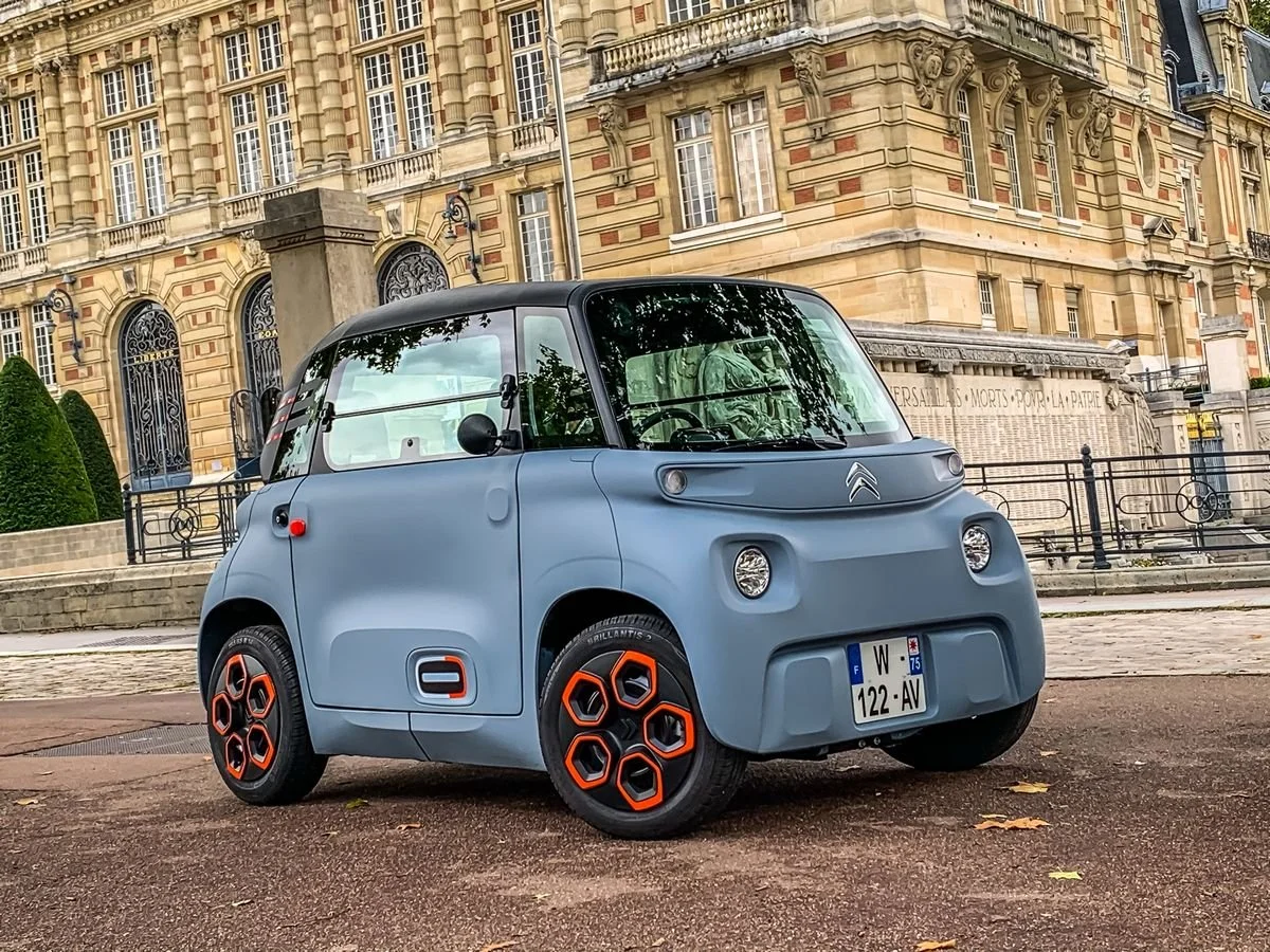Une petite voiture électrique grise avec des détails noirs et orange, garée sur la rue devant un bâtiment ancien en pierre. L'arrière-plan montre une façade historique avec fenêtres à meneaux