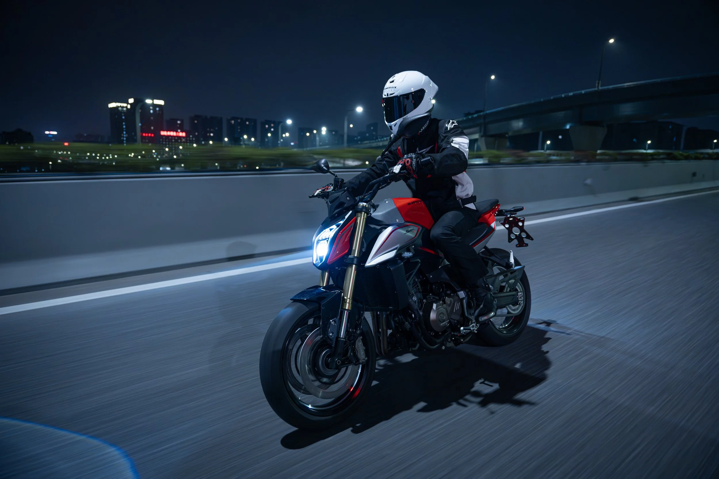 Une personne portant un casque et une veste de moto, chevauchant une moto sportive rouge et noire de nuit sur une autoroute urbaine. Ville en arrière-plan avec bâtiments illuminés.