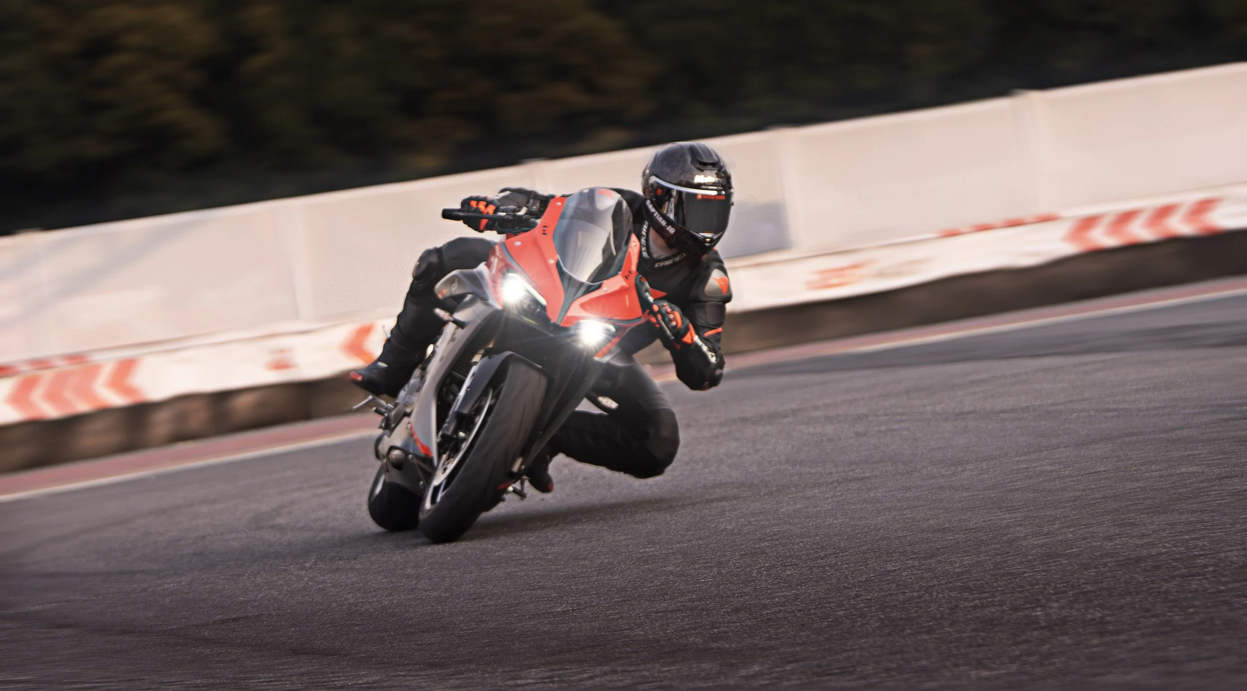 Un motard en tenue noire et casque noir, roulant à grande vitesse dans un virage sur une voie asphaltée, avec un mur de sécurité blanc en arrière-plan.