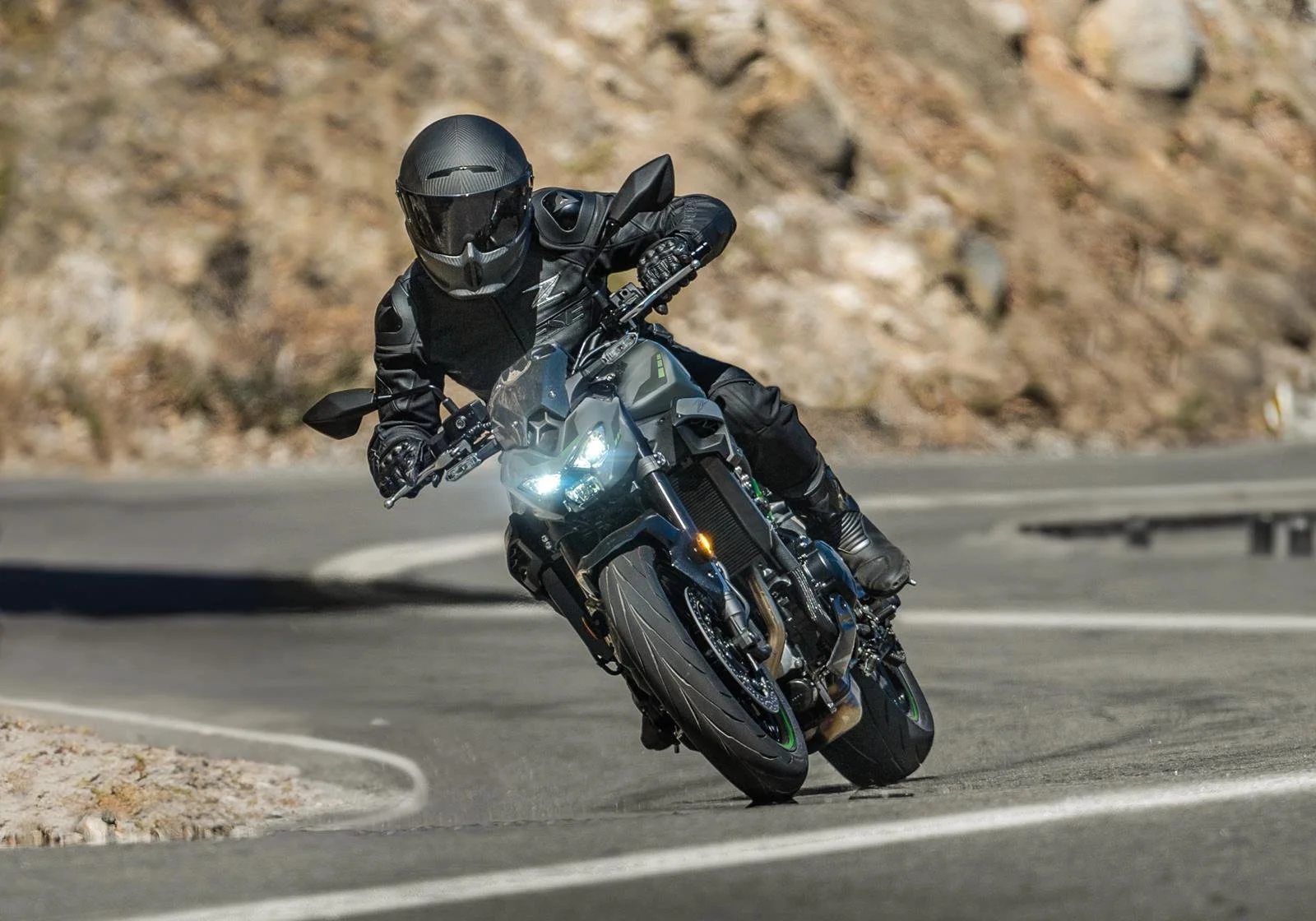Un motard en noir, portant un casque intégral, chemise de cuir, et gants, en train de prendre un virage à grande vitesse sur une route sinueuse en montagne, avec un paysage rocheux en arrière-plan.