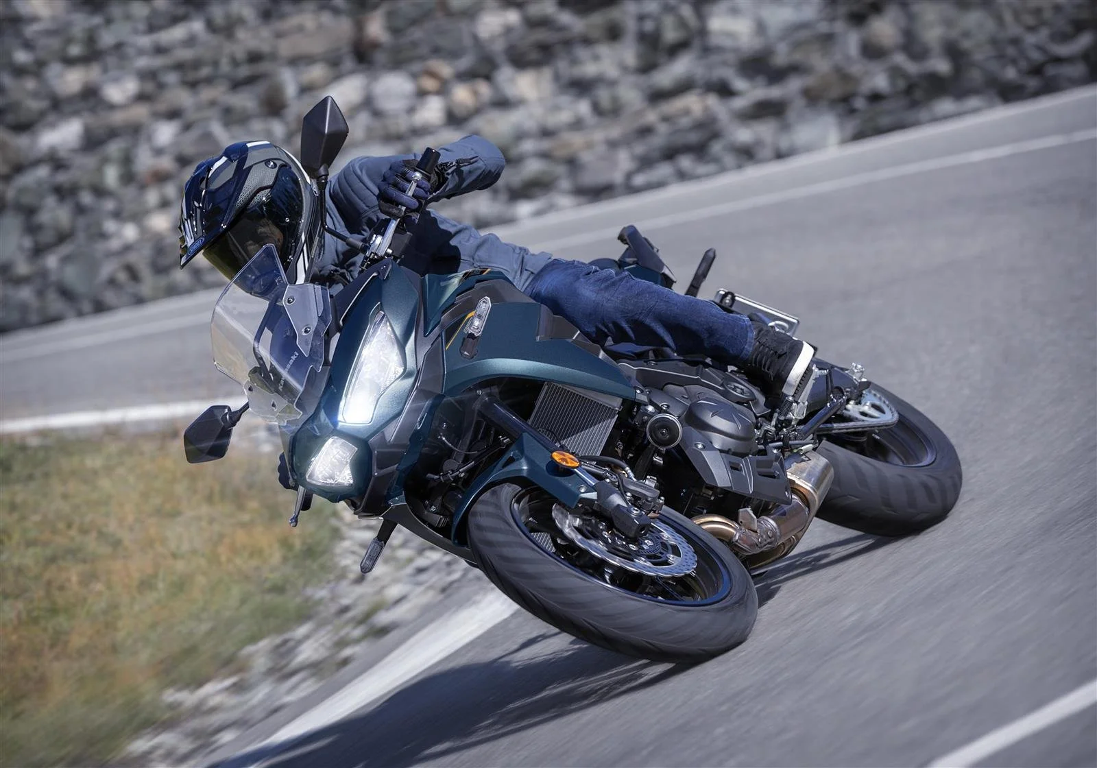 Un motard portant un casque noir et une veste en jean, qui roule à grande vitesse sur une route de montagne en courbe, à moto noire et bleue.